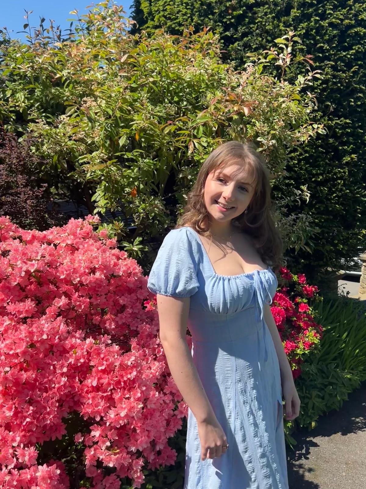 Emily, a young white woman with wavy brown hair wearing a pale blue short-sleeved dress, is standing in the sunlight in front of bushes and bright pink flowers and smiling.