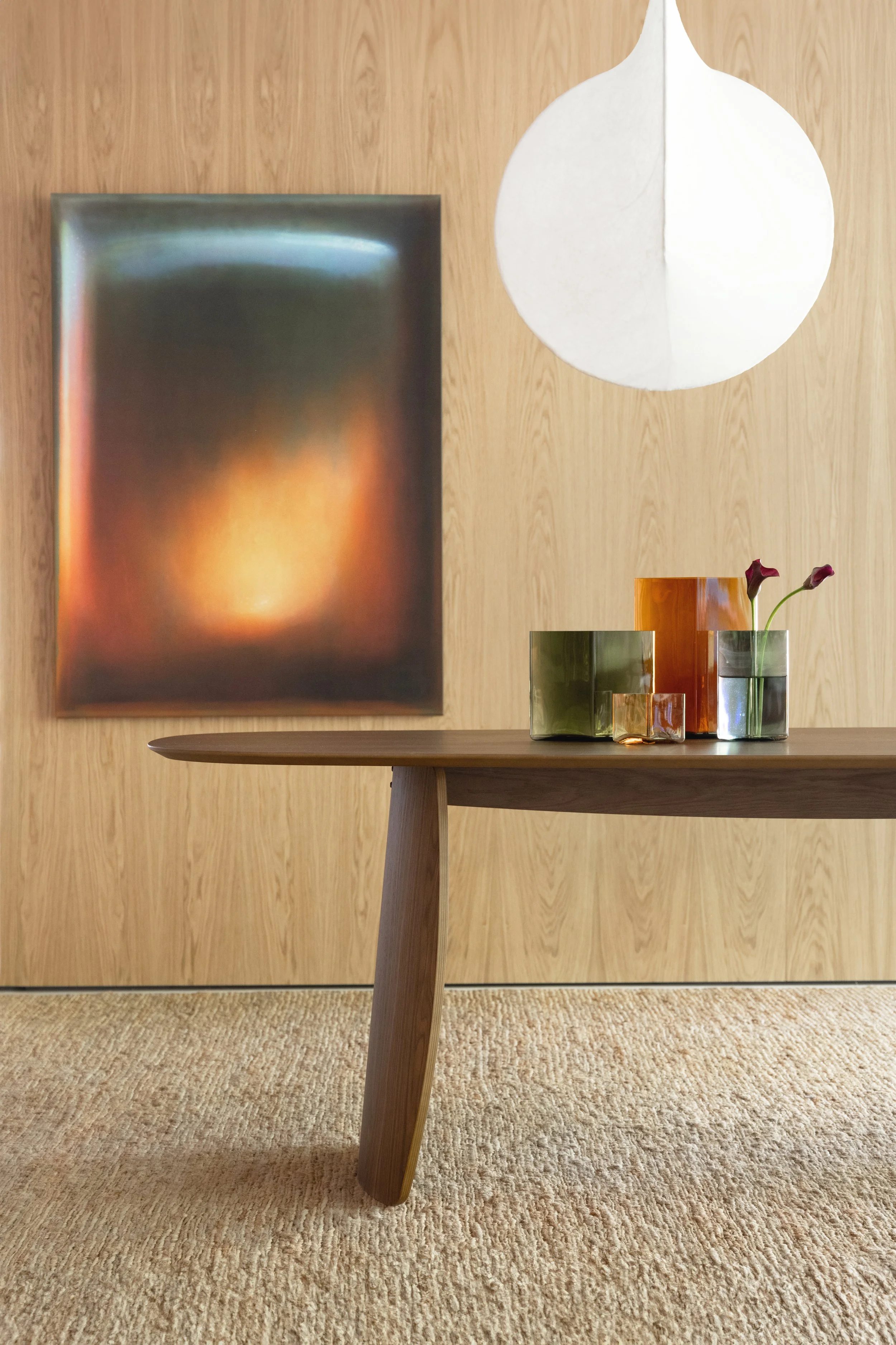 Mesa de madera con flores en vasos de vidrio de colores y una lámpara blanca de diseño moderno en un fondo de pared de madera y alfombra beige.
