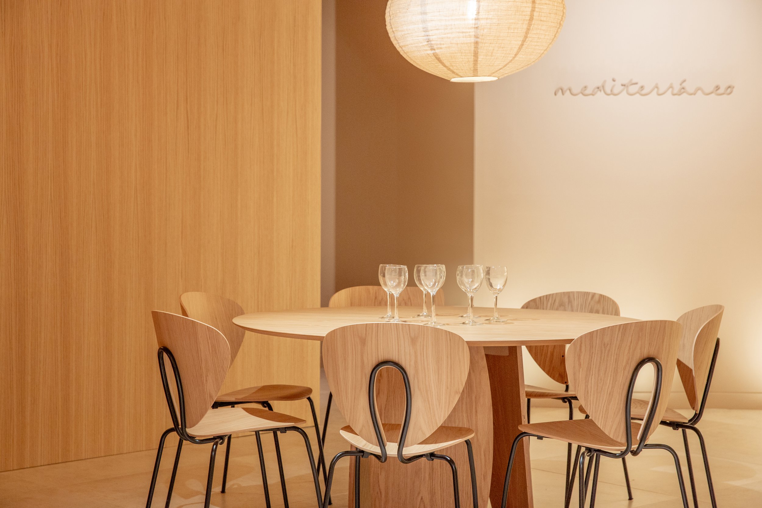 Mesa de madera con siete copas de vino y siete sillas de madera en un restaurante con paredes de madera y decoraciones minimalistas.