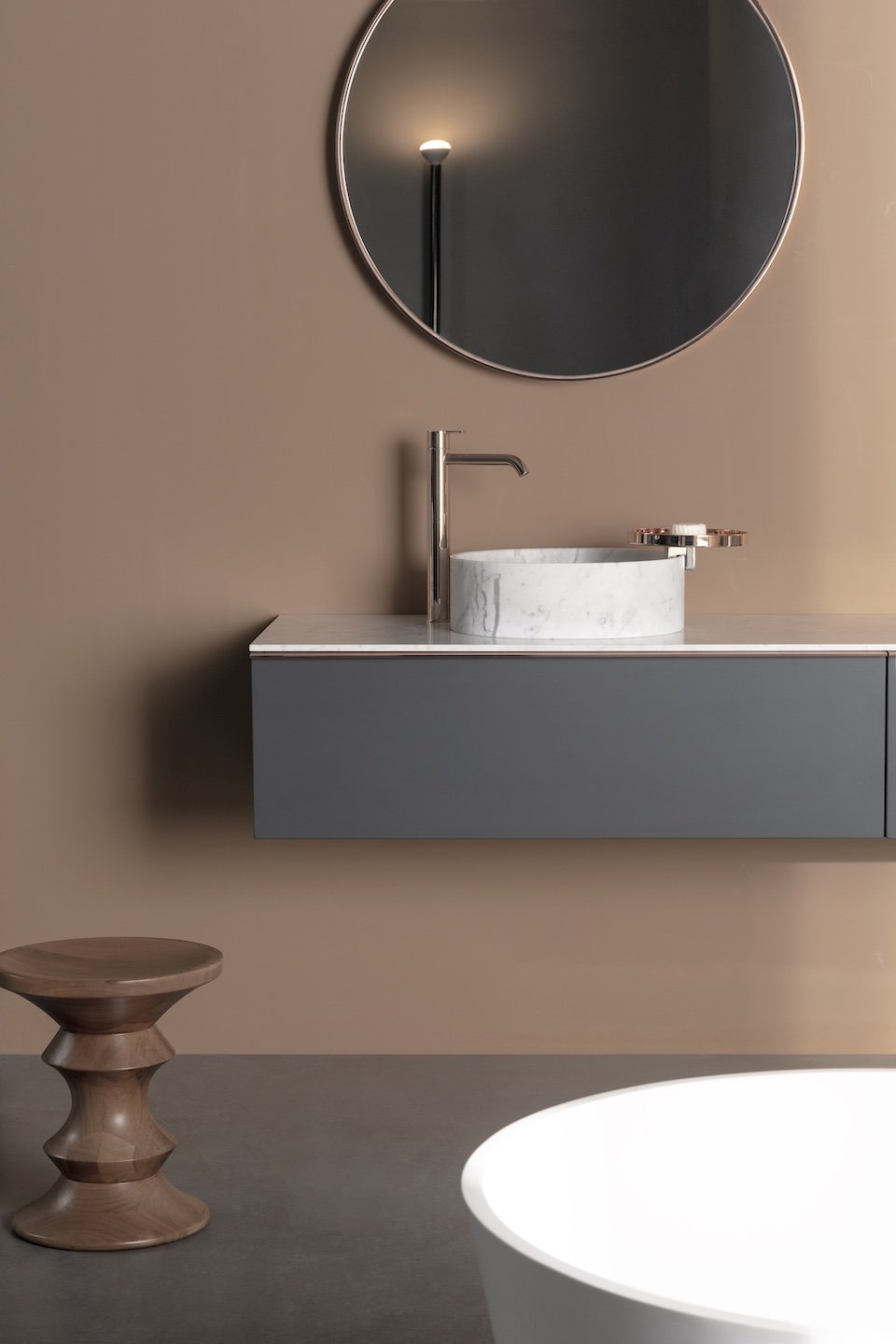 Un lavabo de oro, una mesa de madera con forma de espiral, un lavabo de mármol ovalado en un mueble gris, un espejo redondo y una lámpara de pie sobre una pared de color beige.