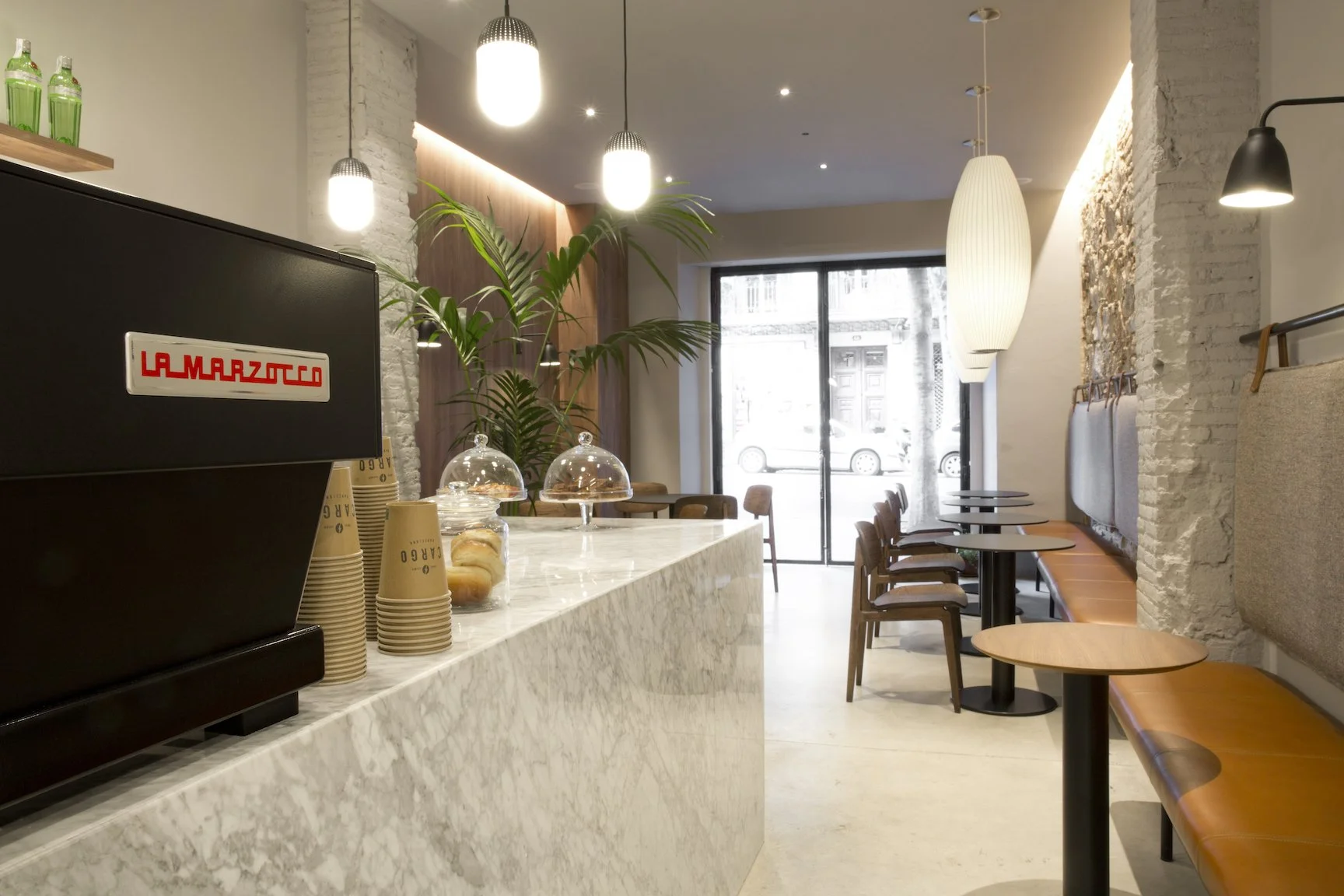 Interior de una cafetería moderna con mesas de madera, sillas de madera, paredes de ladrillo pintadas de blanco, lámparas colgantes y una vitrina con dulces sobre una superficie de mármol blanco.