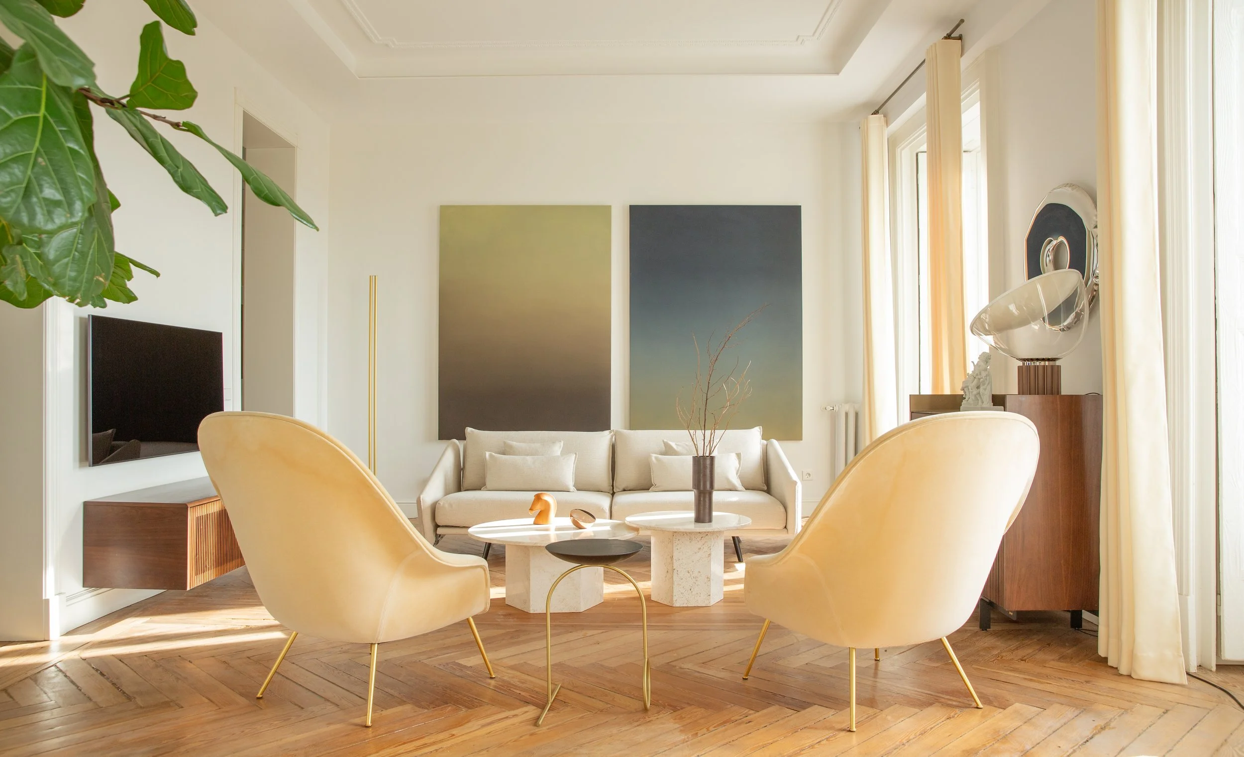 Sala de estar con sillones color crema, mesa central, cortinas claras, cuadros abstractos en la pared, planta grande y decoración moderna.