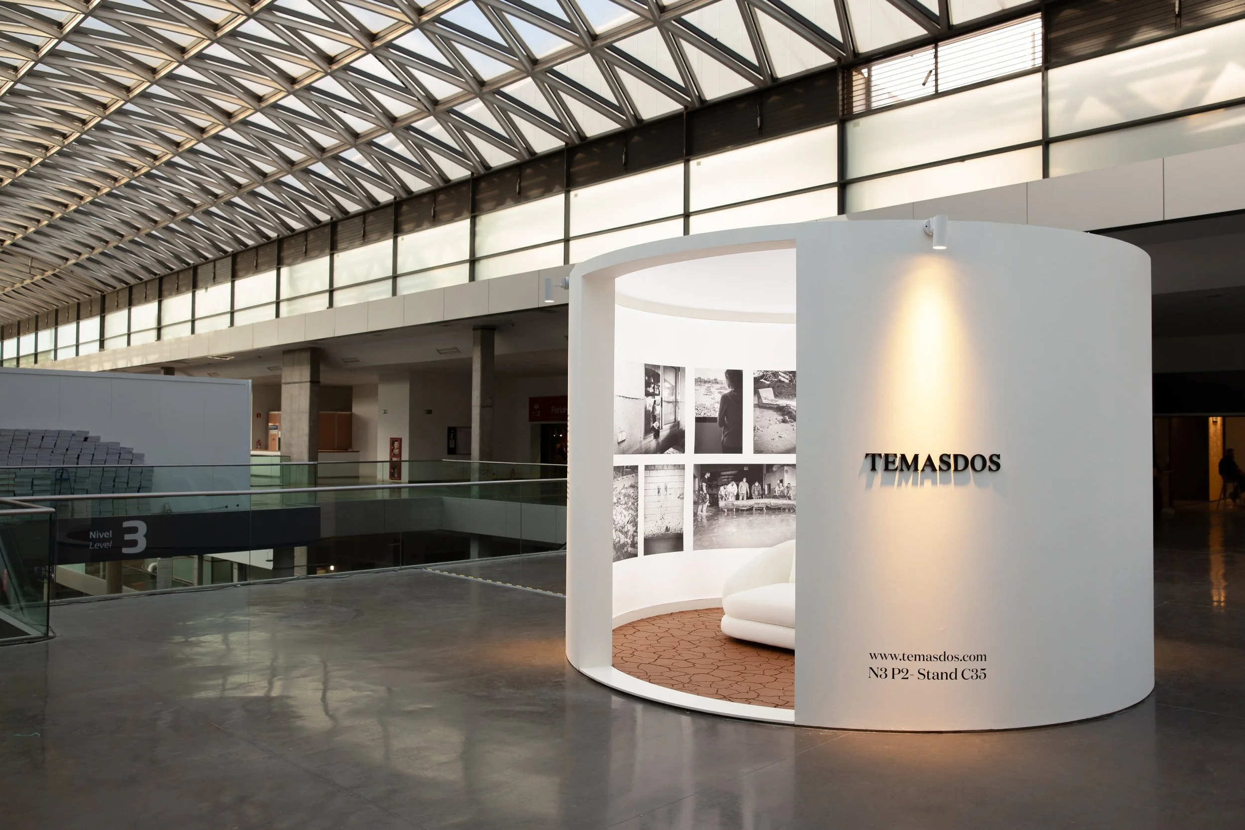 Mostrador de exhibición con cartel que dice 'TEMASDOS' y muestra fotos en blanco y negro en una zona interior de un edificio moderno con techo de vidrio y estructura de metal.