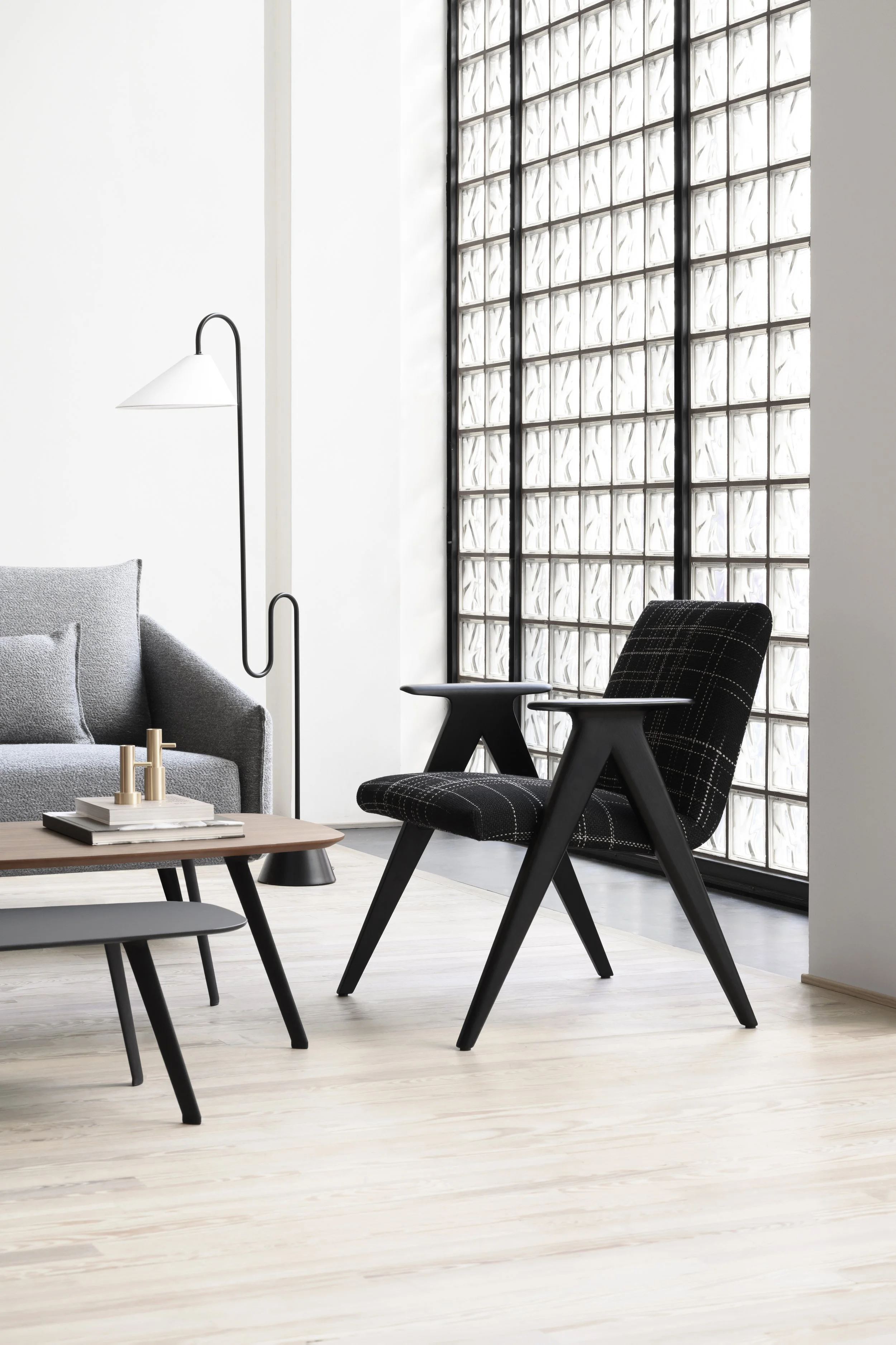 Un espacio de sala moderna con muebles de estilo minimalista. Incluye sillón en gris, una silla de color negro con tapizado de cuadros, mesas de centro con adornos y una lámpara de piso con estructura negra y pantalla blanca. La pared tiene un muro de bloques de vidrio que permite la entrada de luz natural.