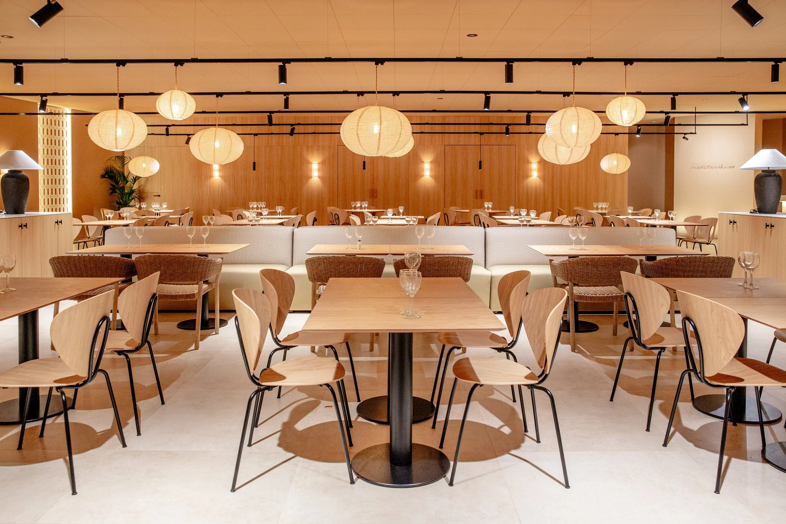 Interior de un restaurante con mesas y sillas de madera, luminarias de papel en el techo y paredes de madera, ambiente moderno y cálido.