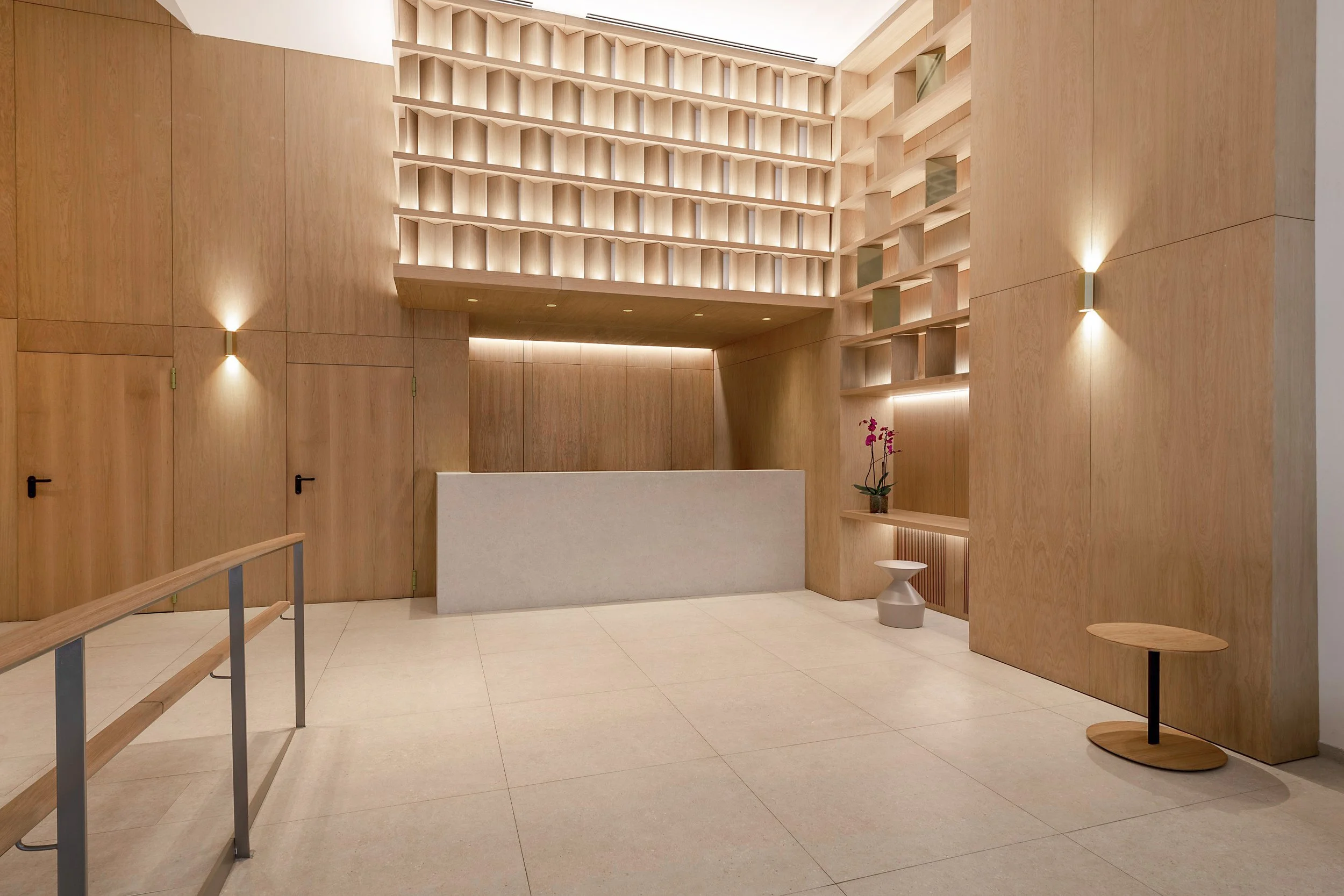 Interior moderno con paredes de madera, estanterías iluminadas y una recepción blanca. Hay un florero con flores y mobiliario minimalista.