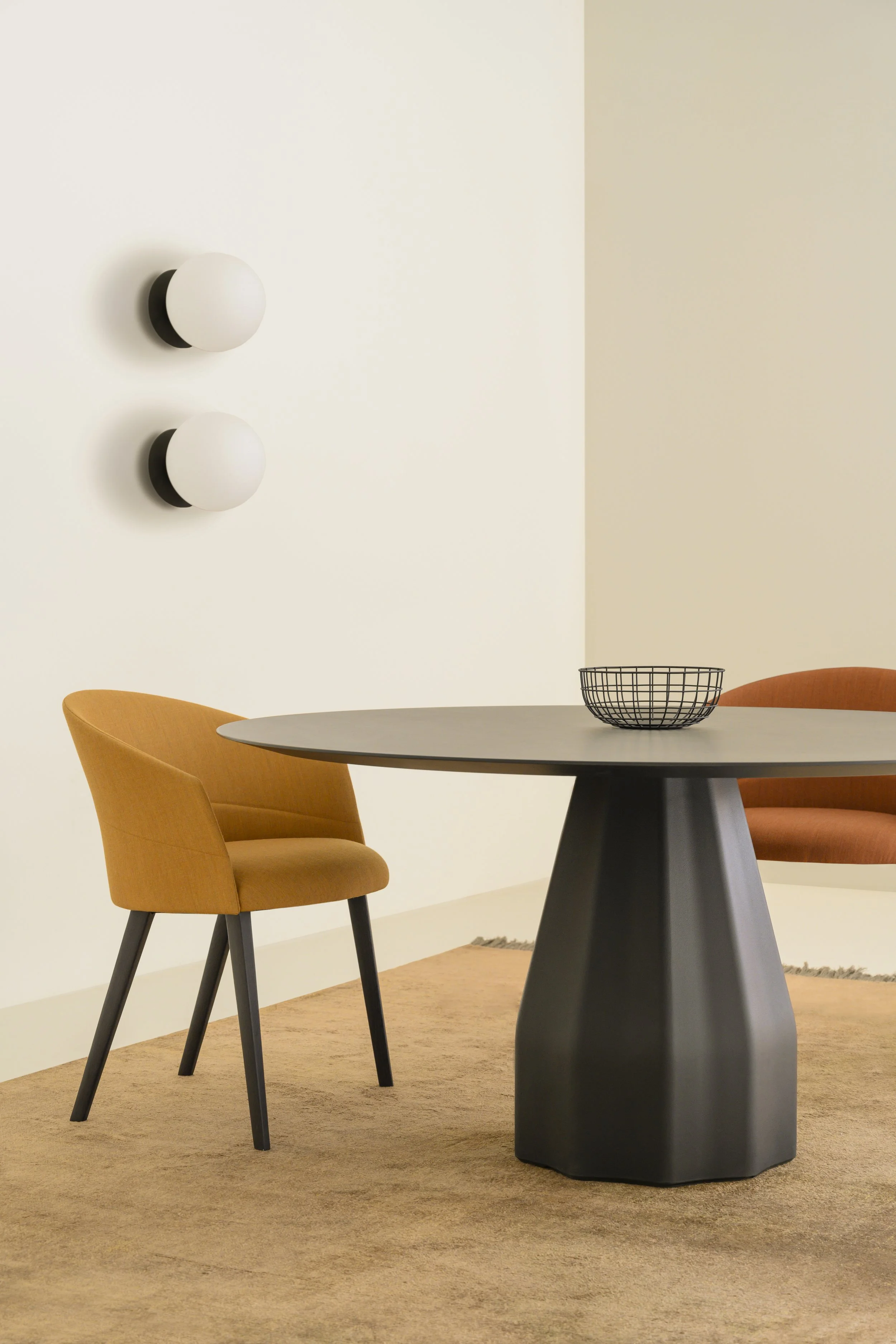 Una mesa redonda de color gris oscuro con una base negra, rodeada de dos sillas de color mostaza con patas negras. Sobre la mesa hay un cesto de alambre negro. En la pared de fondo hay dos lámparas de forma esférica, una en blanco y otra en negro, colocadas una encima de la otra.