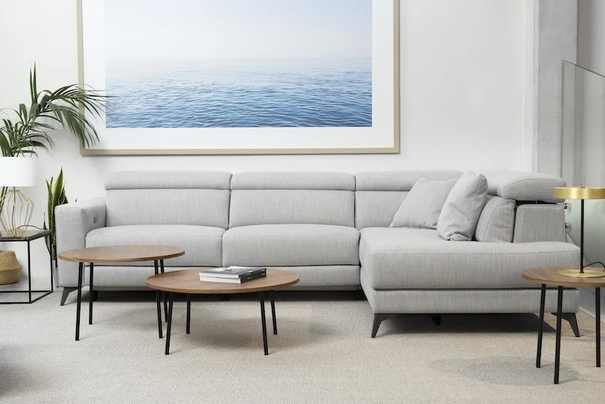 Sala moderna con sofá gris claro, mesas de madera y plantas decorativas, vista de un cuadro con vista al mar en la pared.