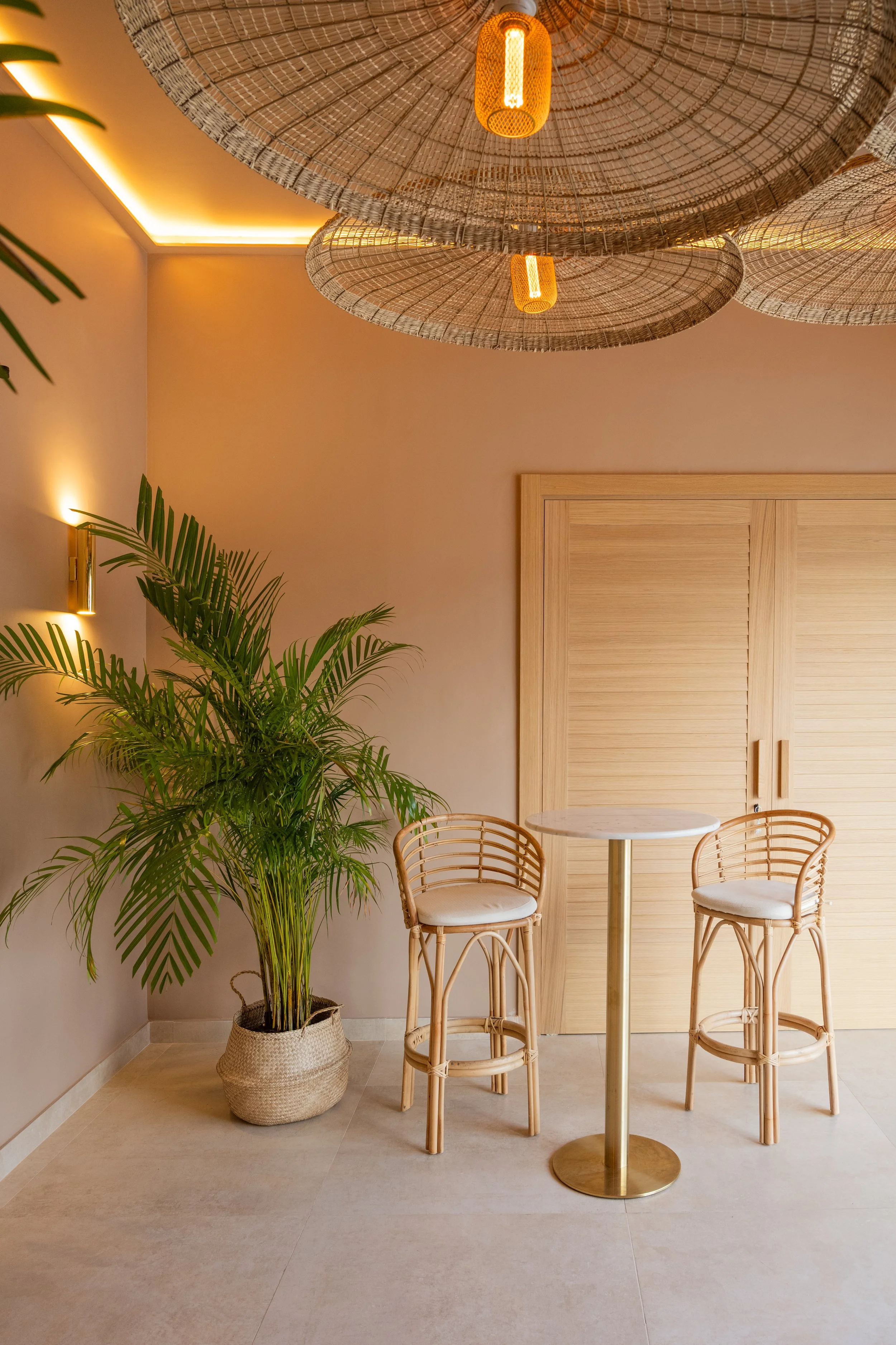 Espacio interior con mesa y sillas de mimbre, planta verde en maceta y lámparas de ratán en el techo, decoración cálida y moderna.