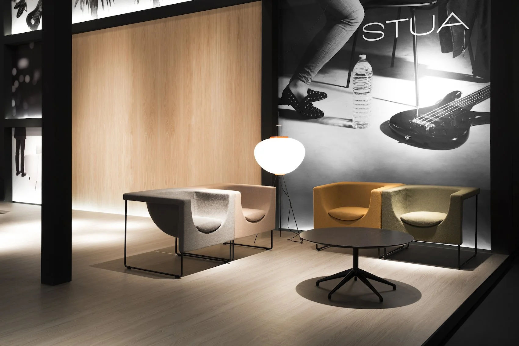 Sala de espera moderna con tres sillones, una lámpara de piso y una mesa redonda de centro, fondo con fotografía de un pie con zapatillas, botella de agua y guitarra eléctrica, en un ambiente acogedor y bien iluminado.