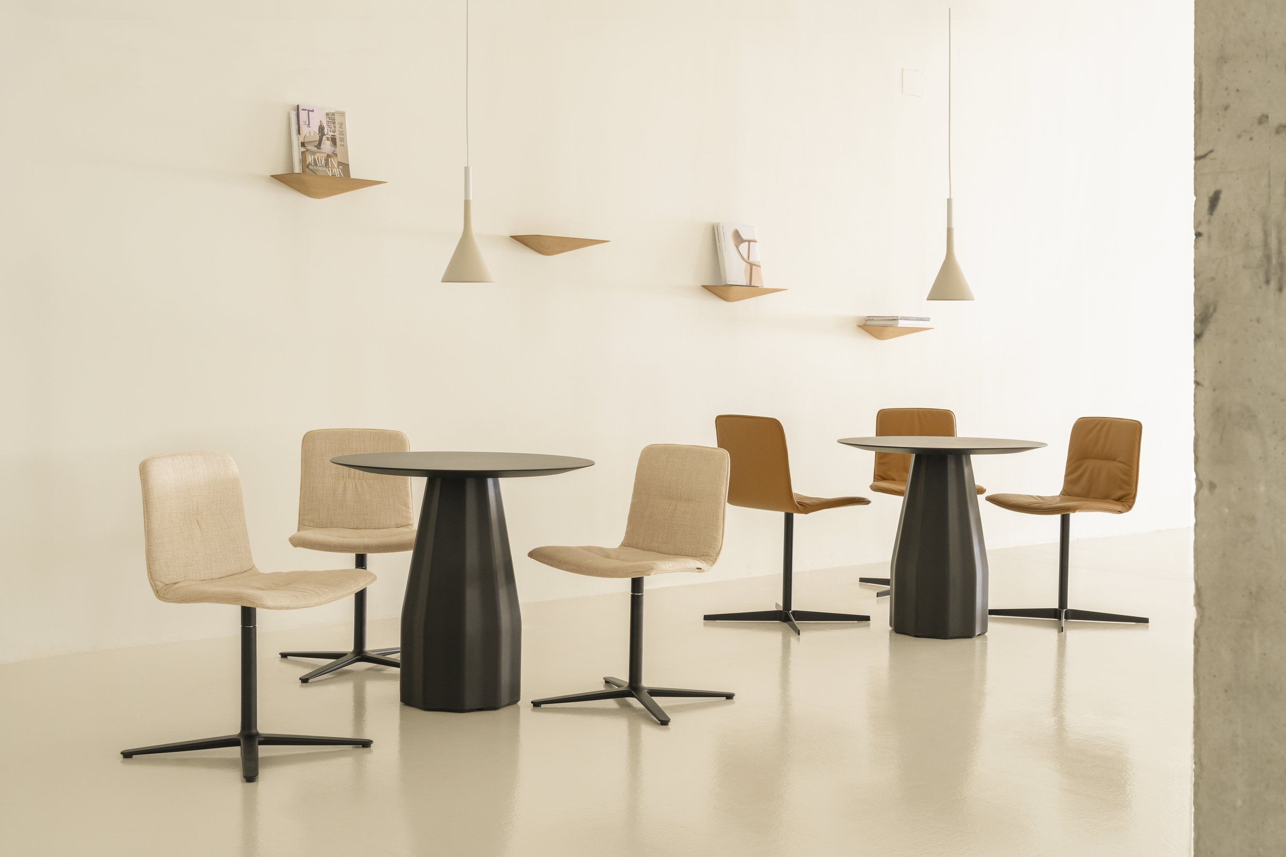 Sala con cuatro mesas redondas y ocho sillas beige y marrón, pared blanca con estantes con libros y lámparas colgantes.