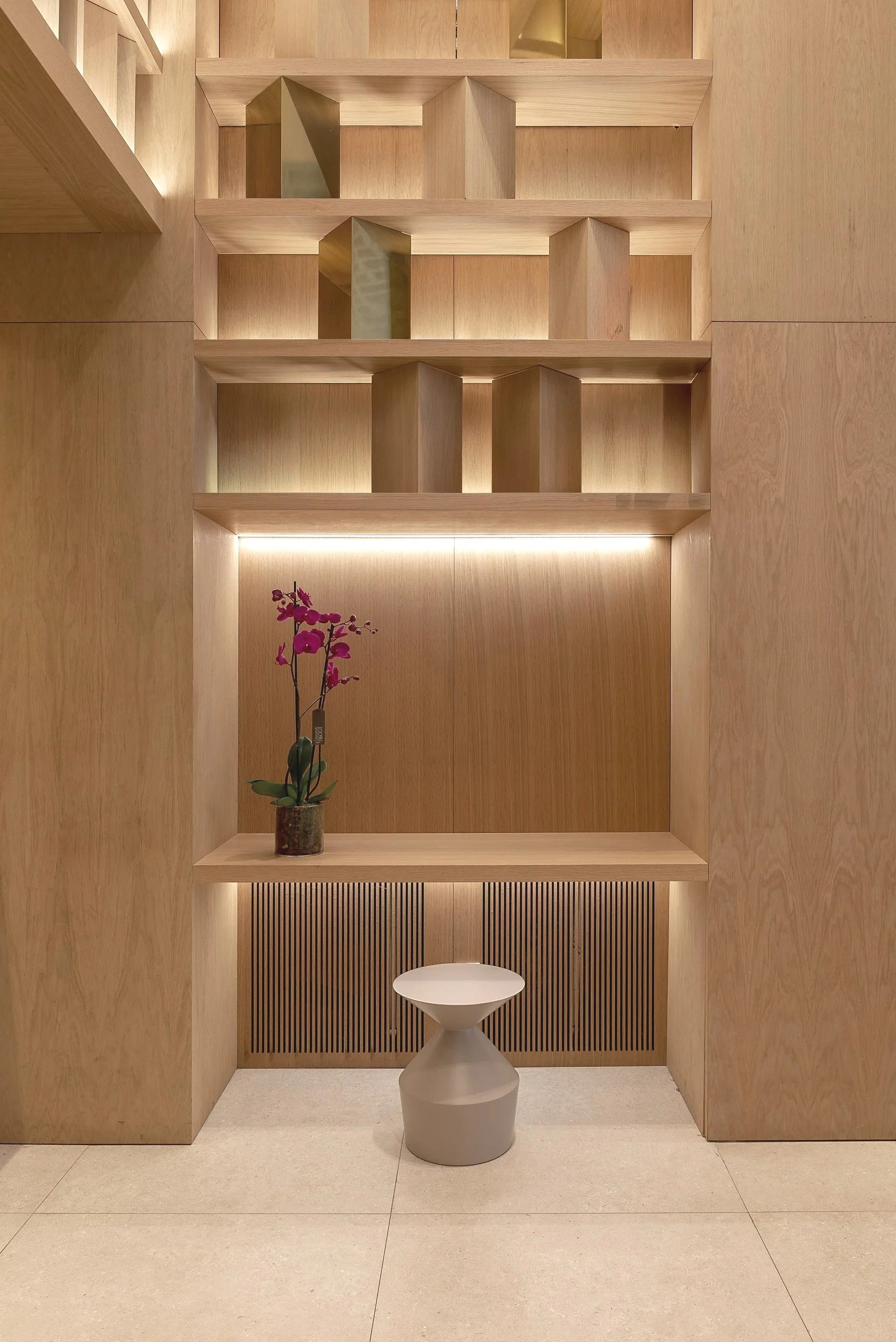 Salón minimalista con paredes de madera, estanterías, una planta de oro de color rosa y un mueble gris con forma de copa en el centro. Hay iluminación suave y un suelo de color claro.