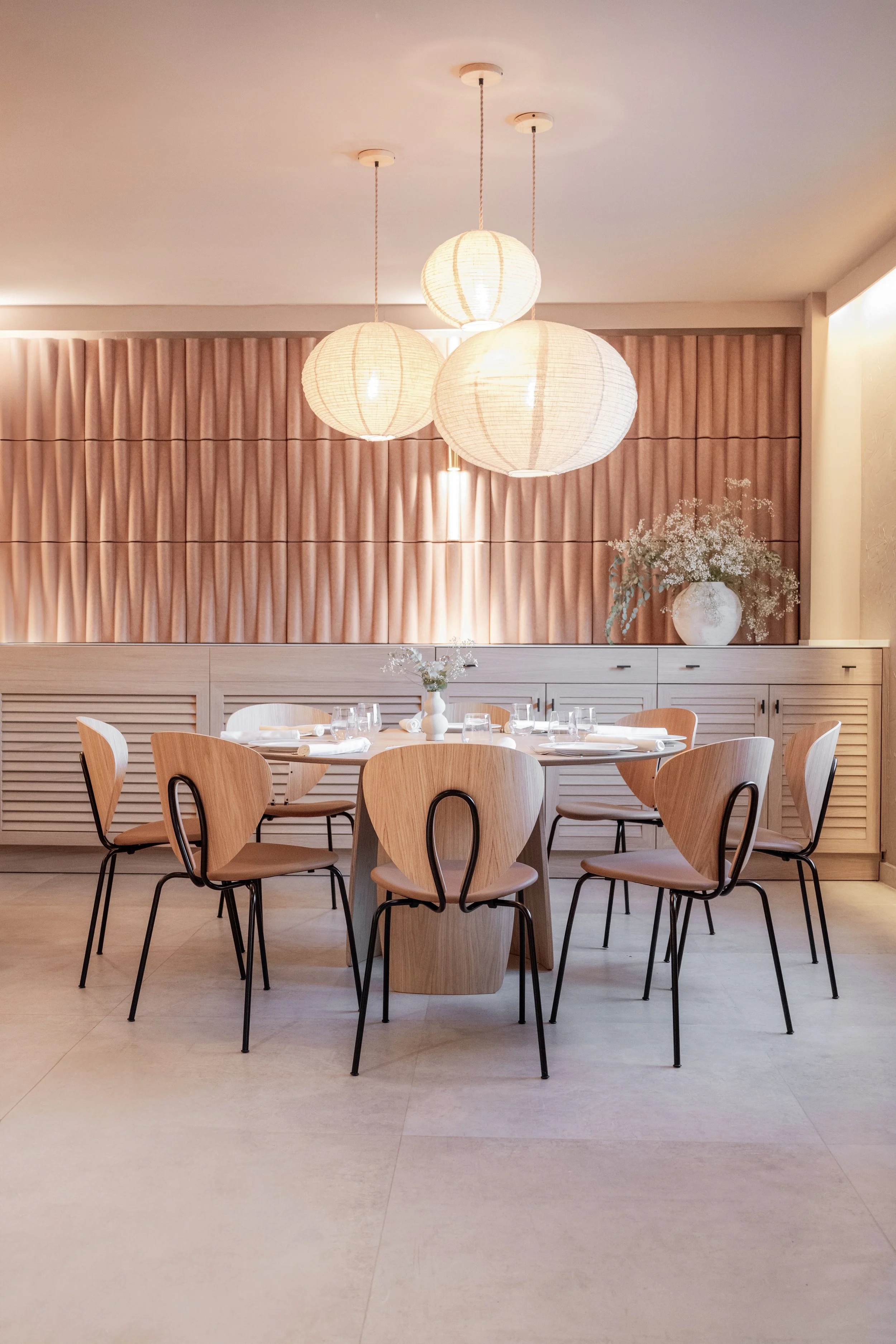Comedor con mesa redonda de madera y sillas de respaldo abierto, decorado con flores blancas en jarrón y colgado de techos con lámparas de papel redondas y candelabros, paredes con paneles de madera y suelo de baldosas claras.