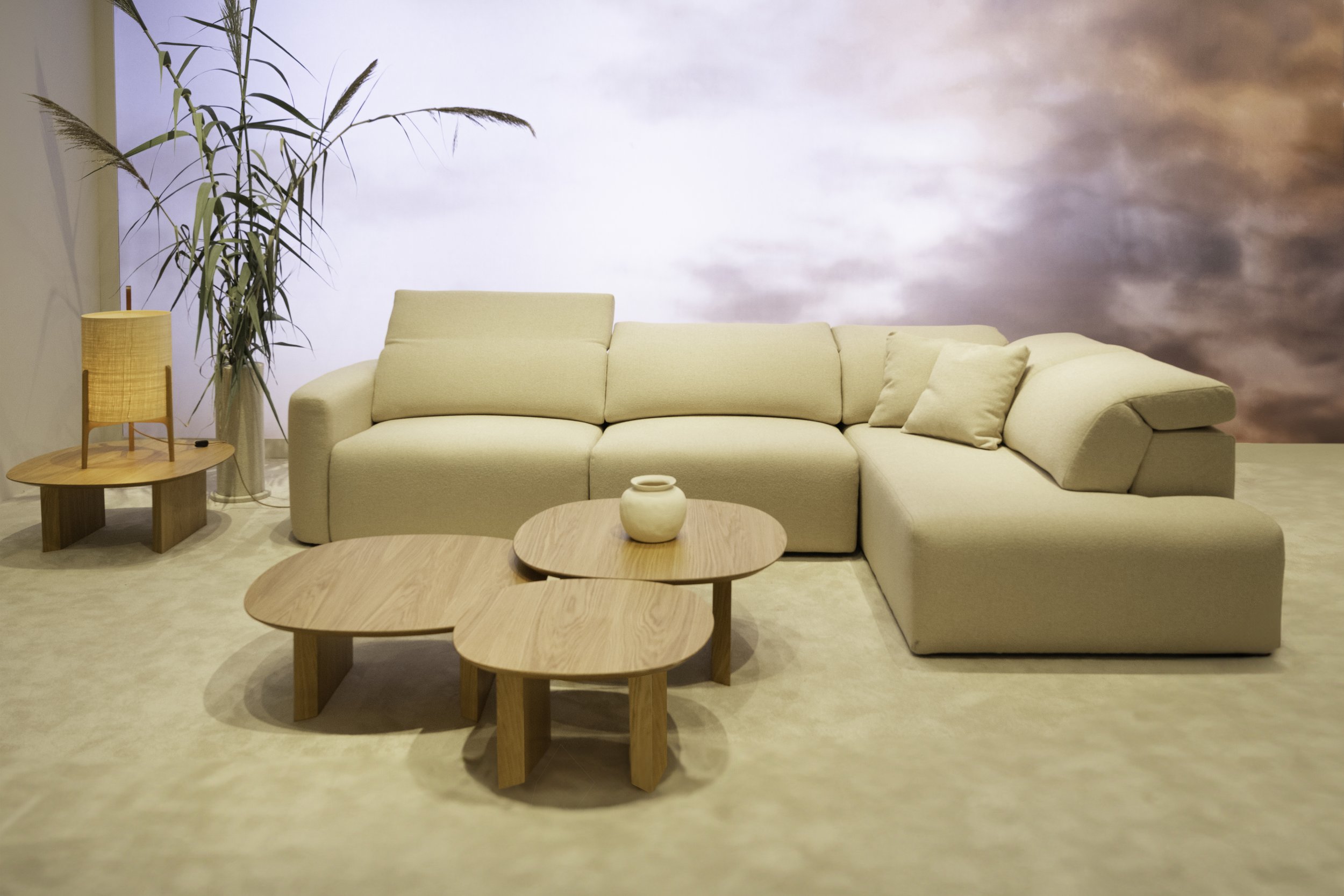 Sala de estar con sofá color crema, mesa de madera, lámpara de pie de tono similar, plantas altas y un fondo decorativo con tonos rosados y lilas.