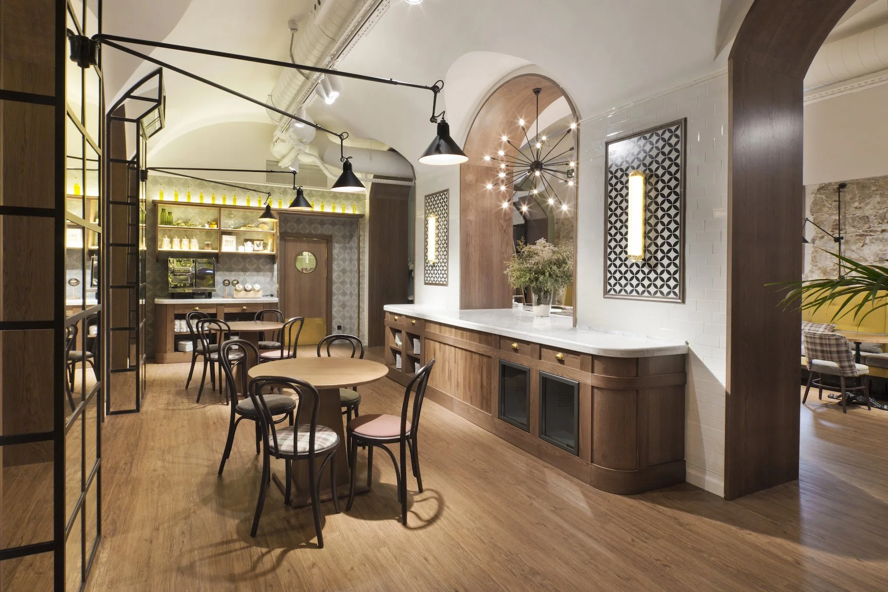 Interiores de cafetería con mesas de madera, sillas de diseño clásico, una barra de madera con encimera de mármol, decoraciones modernas, y luces colgantes, creando un ambiente cálido y acogedor.