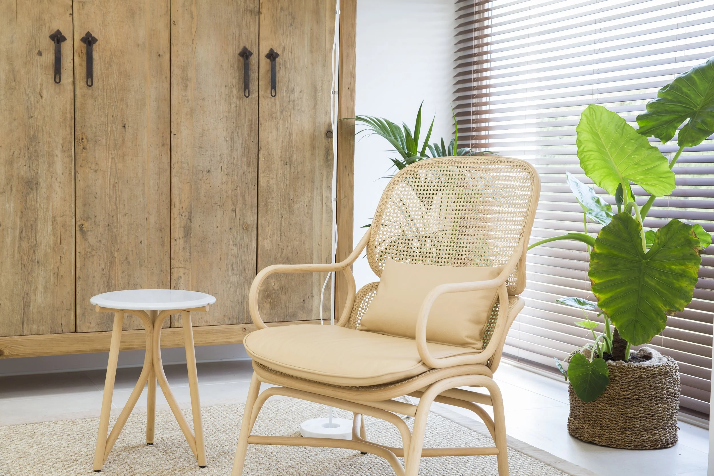 Un rincón de sala con sillón de mimbre, mesa pequeña de madera y plantas verdes junto a ventanas con persianas horizontales.