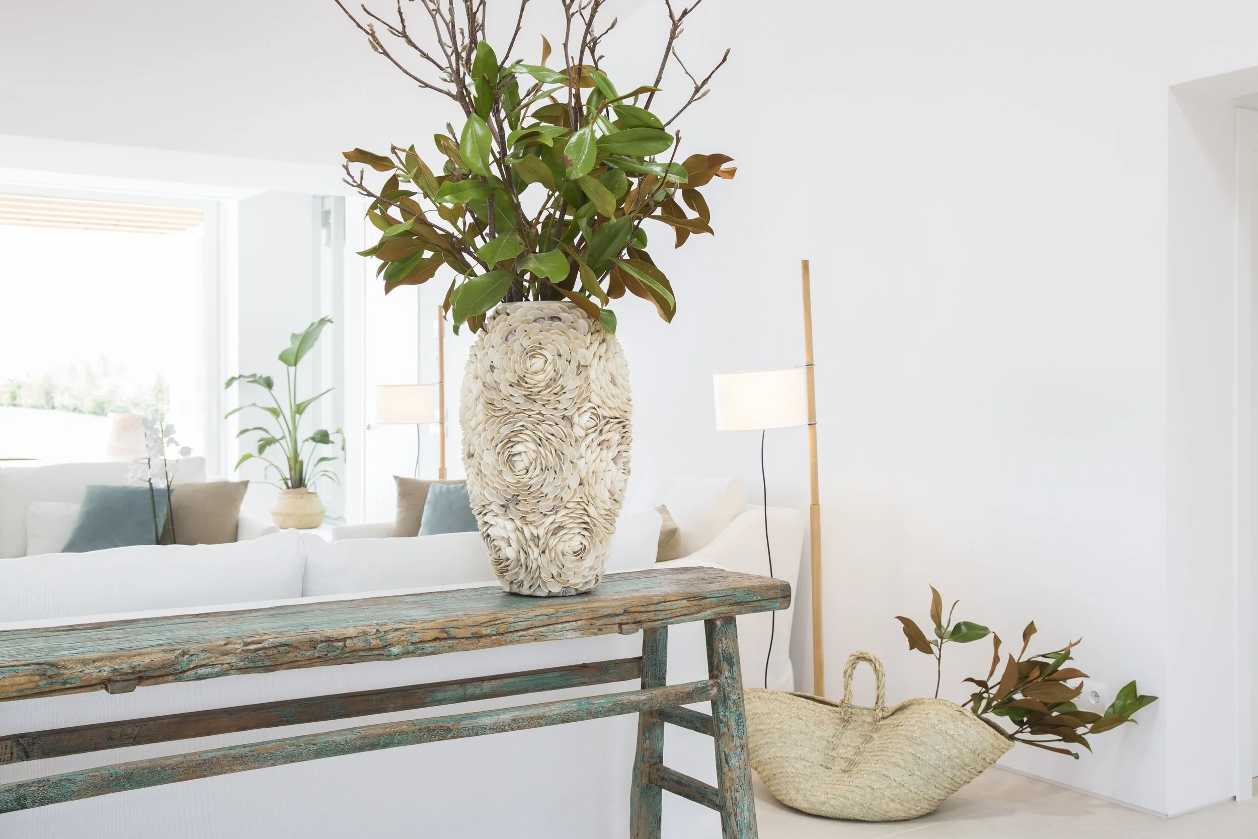 Florero decorativo con ramas y hojas verdes en un espacio interior con muebles blancos y lámparas, en un estilo minimalista y luminoso.