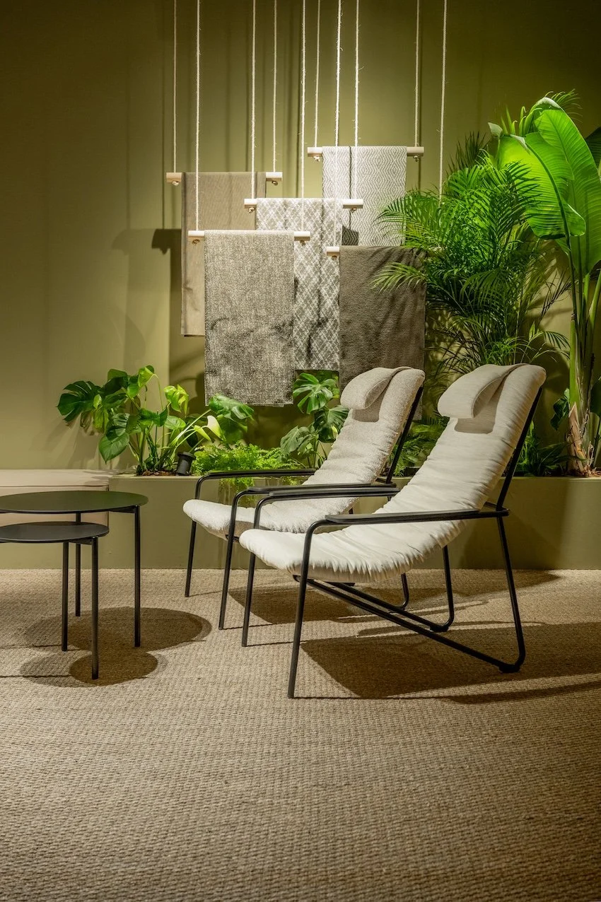 Dos sillas blancas con estructura negra, mesas pequeñas negras, plantas verdes y alfombra beige, con una pared verde de fondo con colgados toallas o telas.