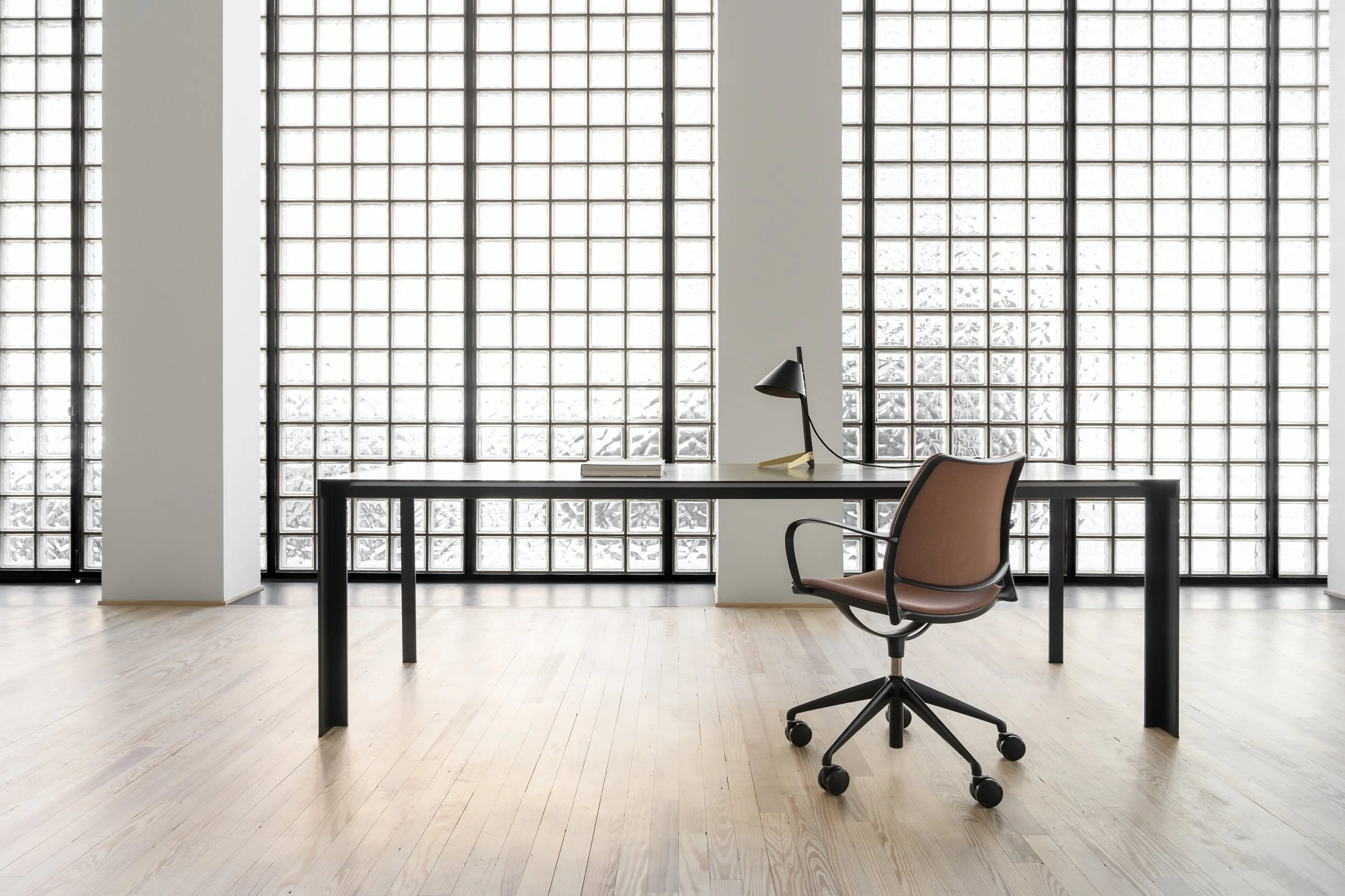 Oficina moderna con muro de bloques de vidrio, mesa de trabajo negra, escritorio con lámpara negra y silla rosa con base negra en el primer plano.