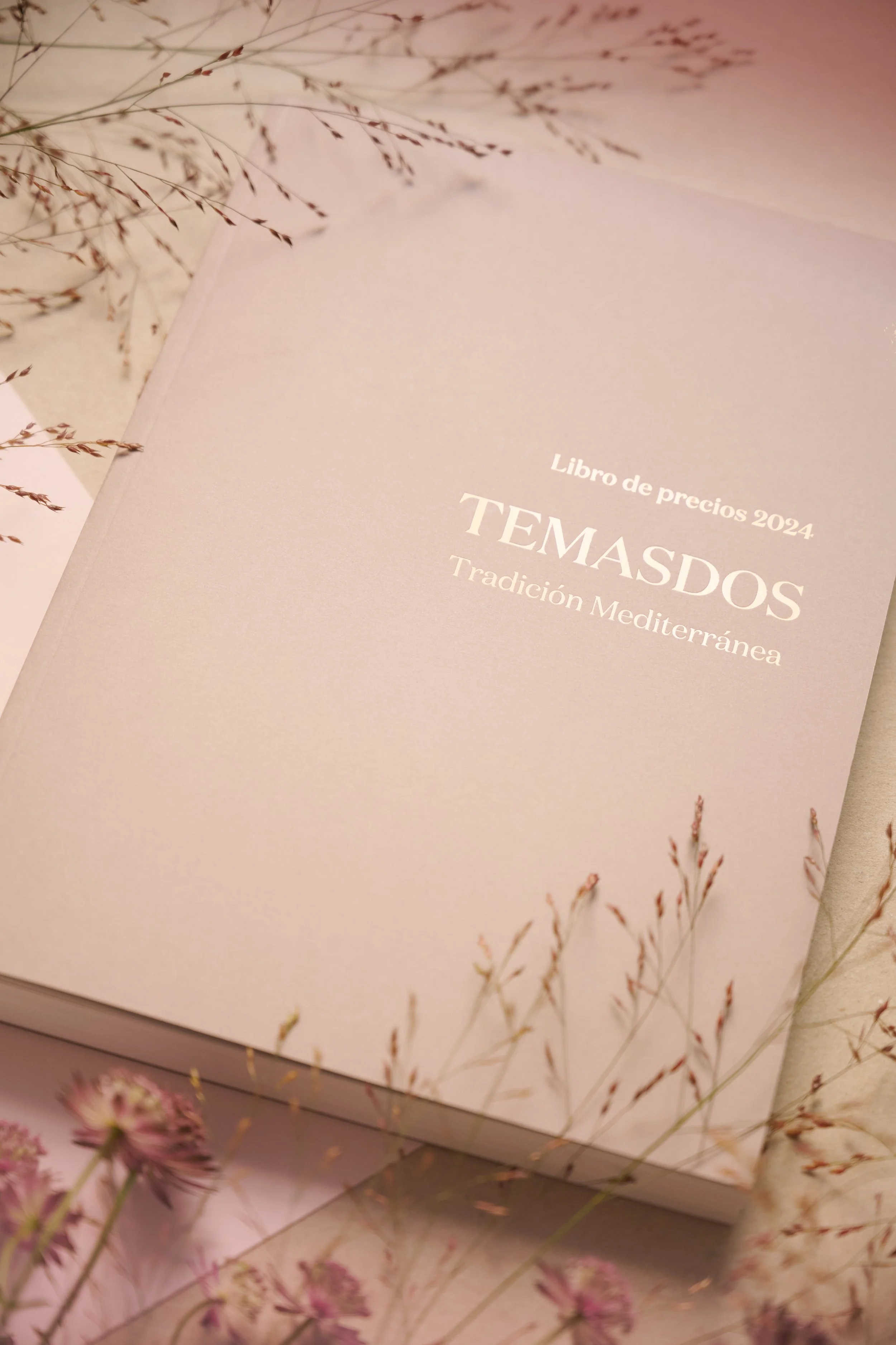 Libro de precios 2024 titulado 'Temasdos, tradición mediterránea', con fondo rosado y detalles de flores secas en la esquina superior izquierda y en la parte inferior