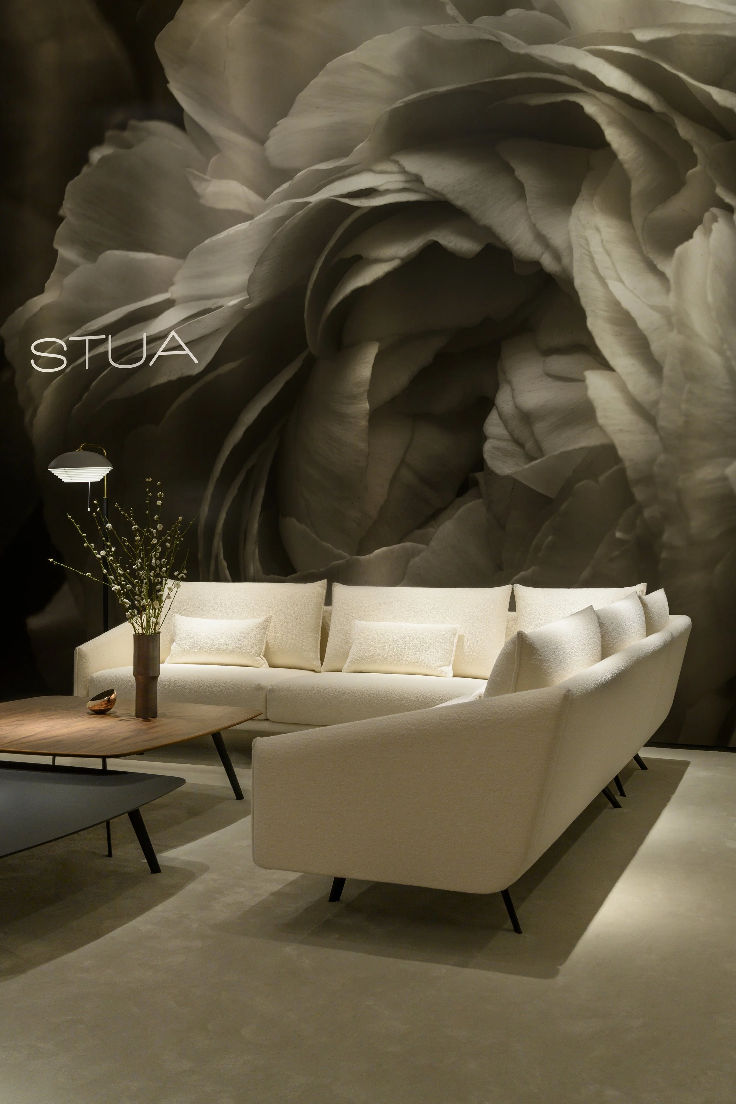Sala de estar moderna con sillones beige, mesa de madera, lámpara de pie y maceta con planta, frente a un gran mural de flores en tonos neutros.