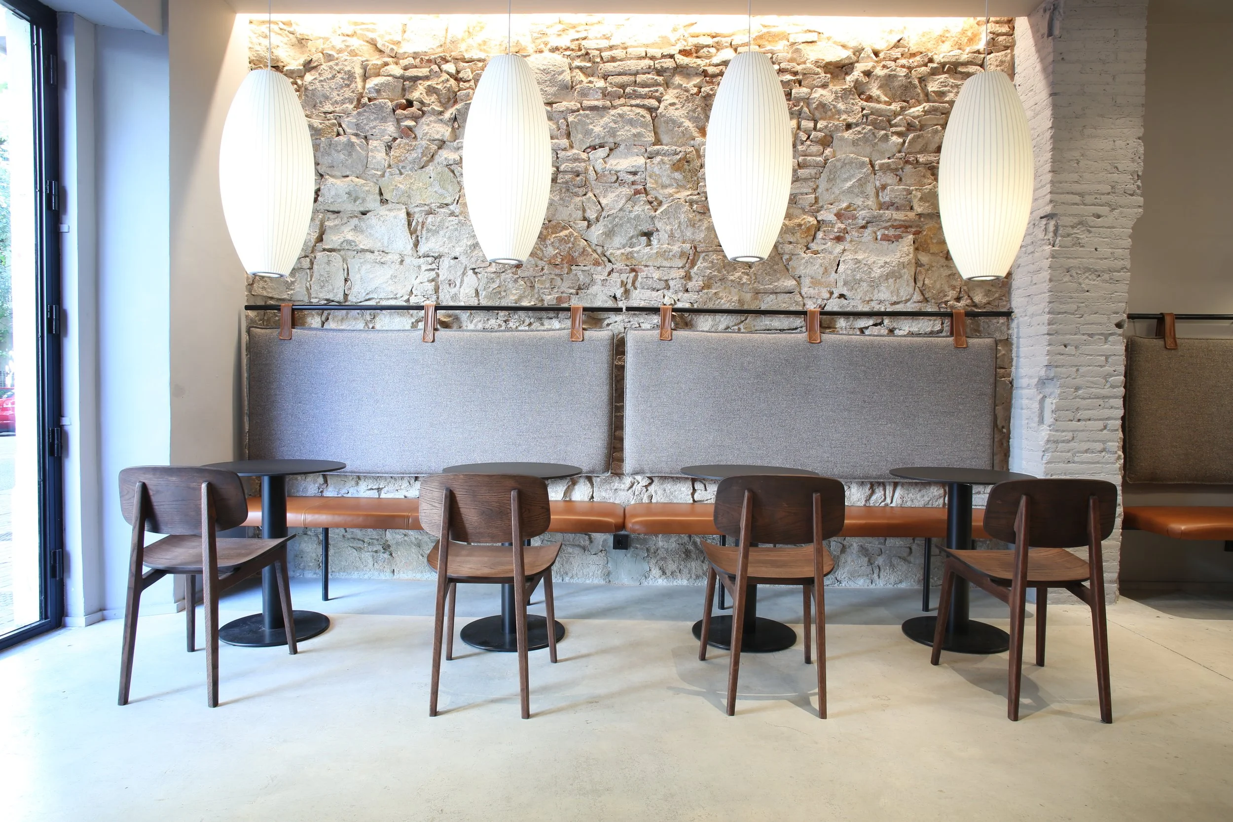 Interiores de un restaurante con paredes de piedra vista, lámparas colgantes blancas y asientos de madera con respaldo gris claro.