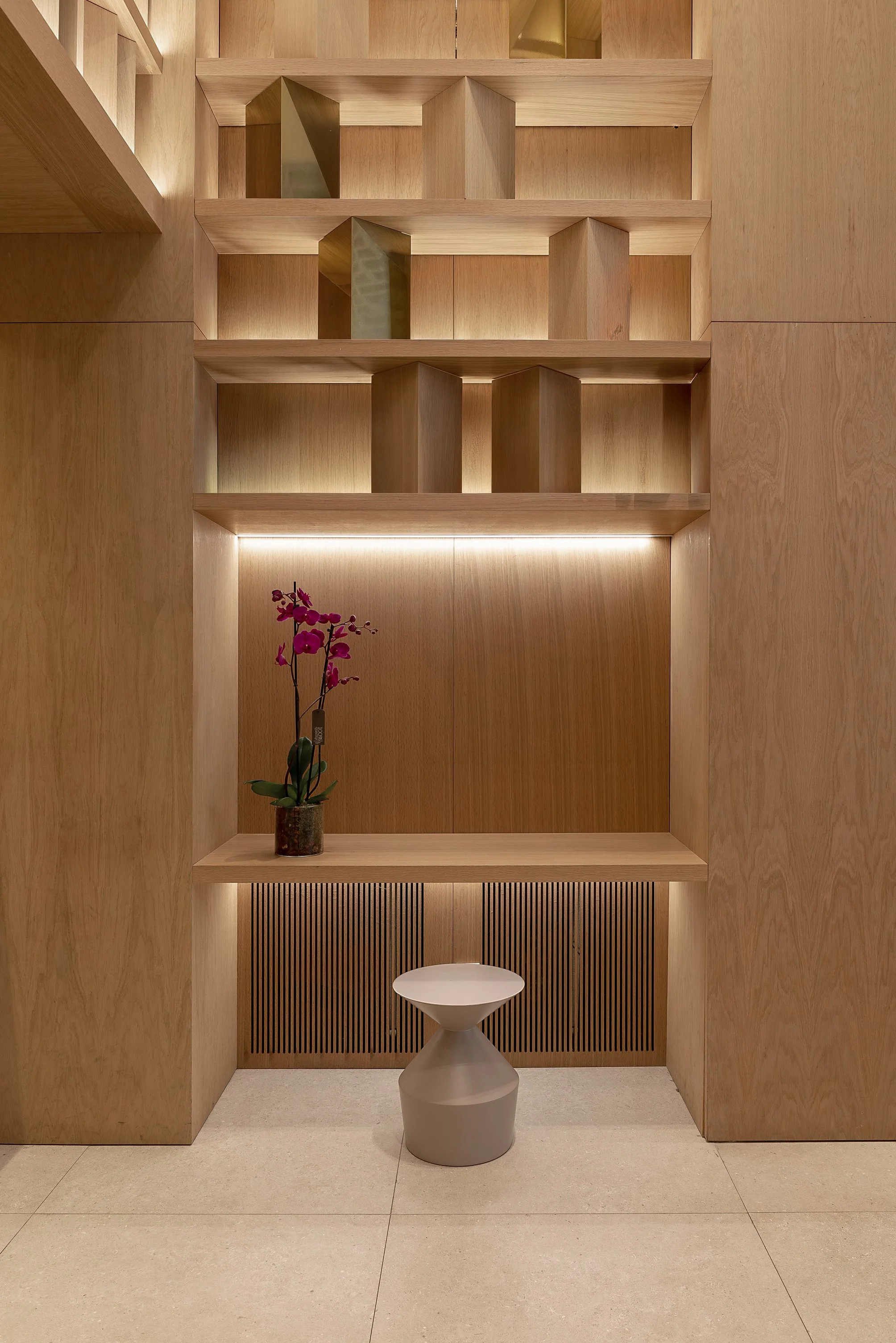 Un espacio minimalista y moderno con paredes y estanterías de madera clara, una planta de orquídea púrpura en una maceta y un taburete de color gris y forma inusual.