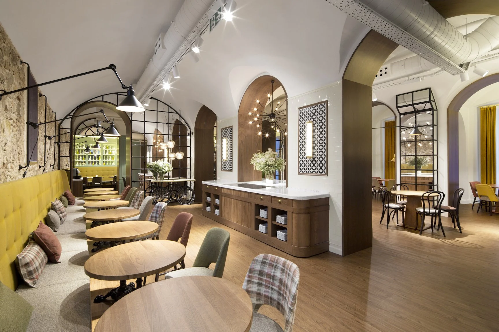 Interior de un restaurante con mesas de madera, sillas tapizadas y decoraciones modernas, arcos y ventilación expuesta en el techo.