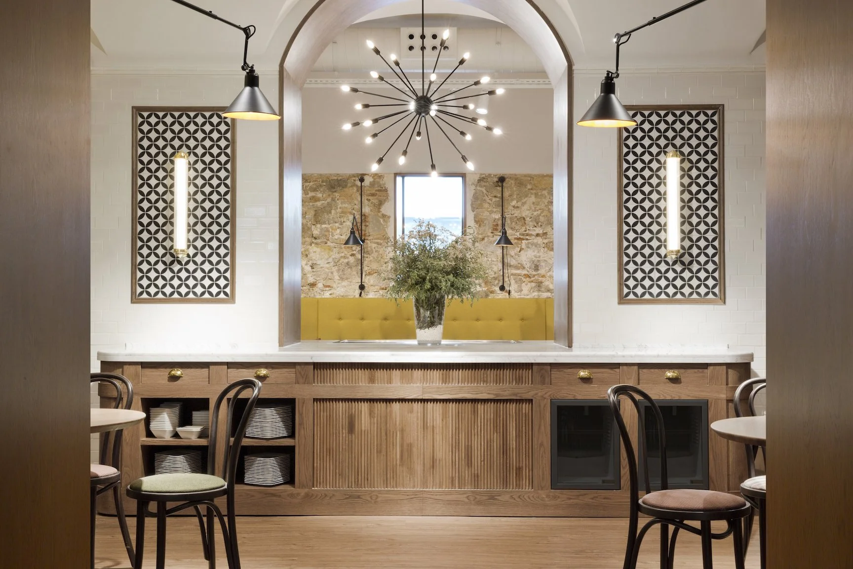 Interior de un restaurante, vista hacia la barra con una planta en un florero, iluminación moderna en el techo, paredes decoradas con paneles de patrones geométricos y ladrillos expuestos en el fondo, mesas con sillas alrededor.