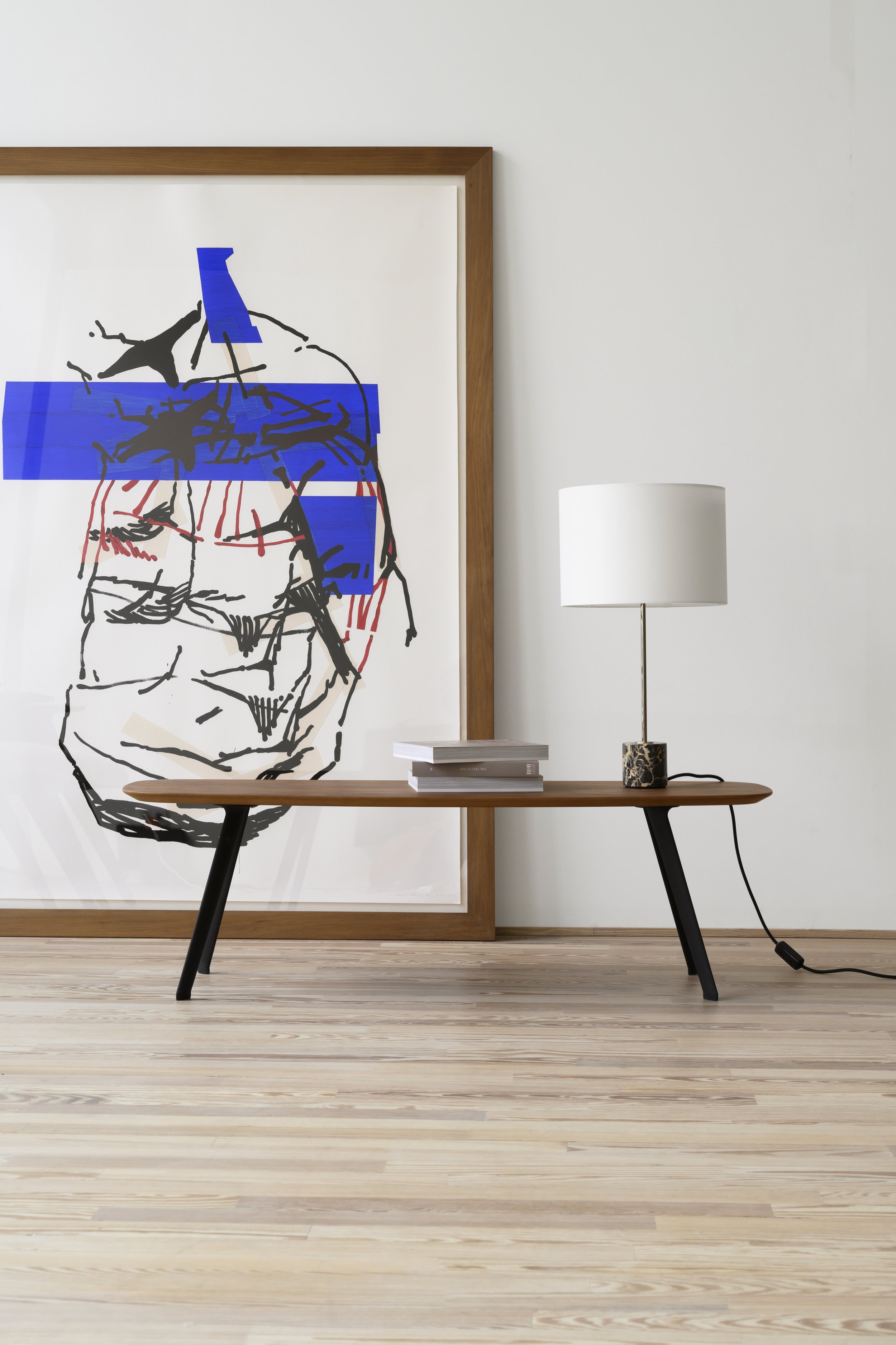 Cuadro con arte abstracto con líneas negras y bloques azules, en un marco de madera, junto a una lámpara con base de mármol y pantalla blanca, sobre una mesa de madera en una sala moderna con paredes blancas y suelo de madera clara.
