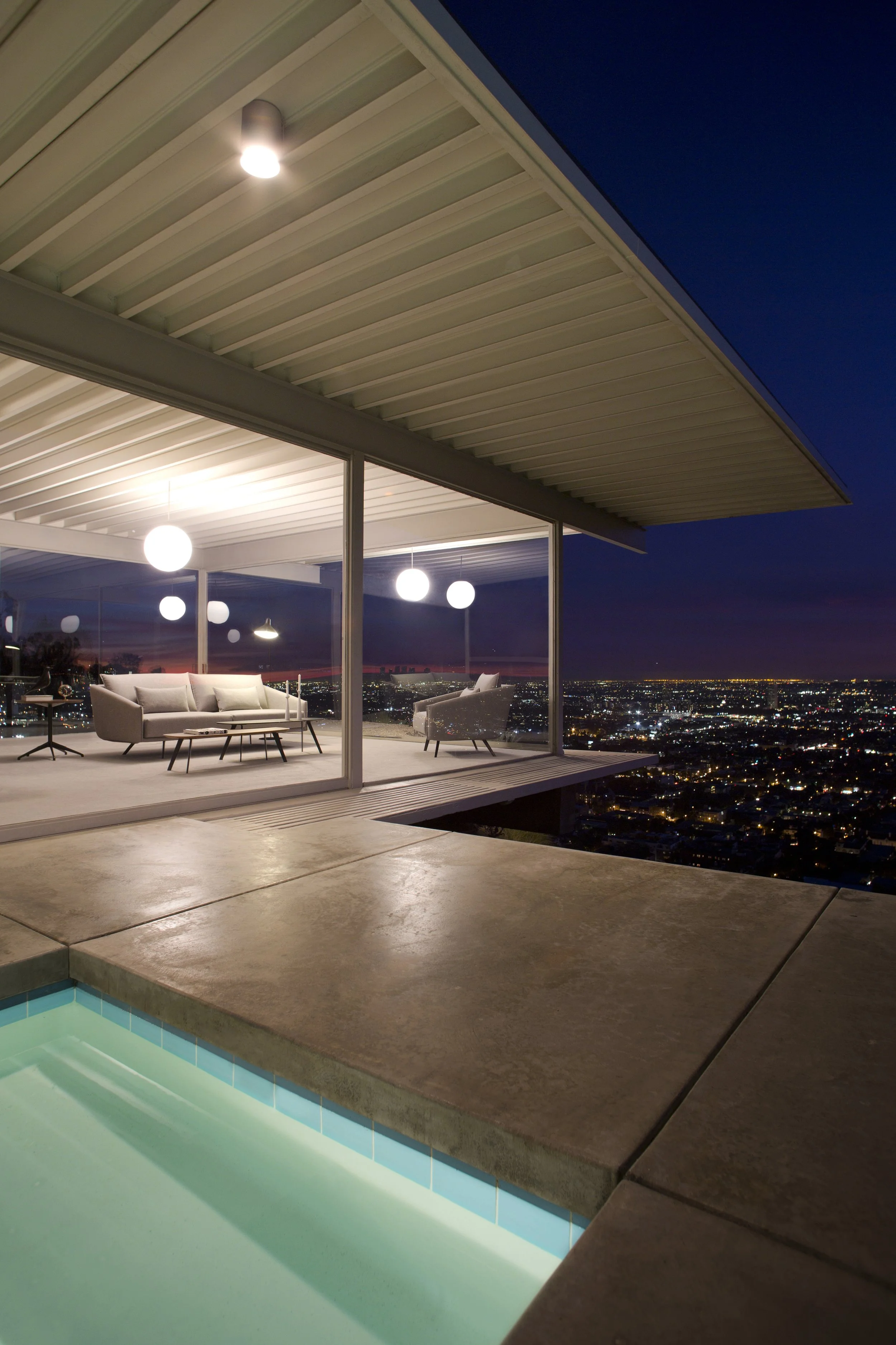 Vista nocturna de terraza moderna con sala de estar, vista a la ciudad iluminada, piscina con agua azul y paisaje urbano en el fondo.