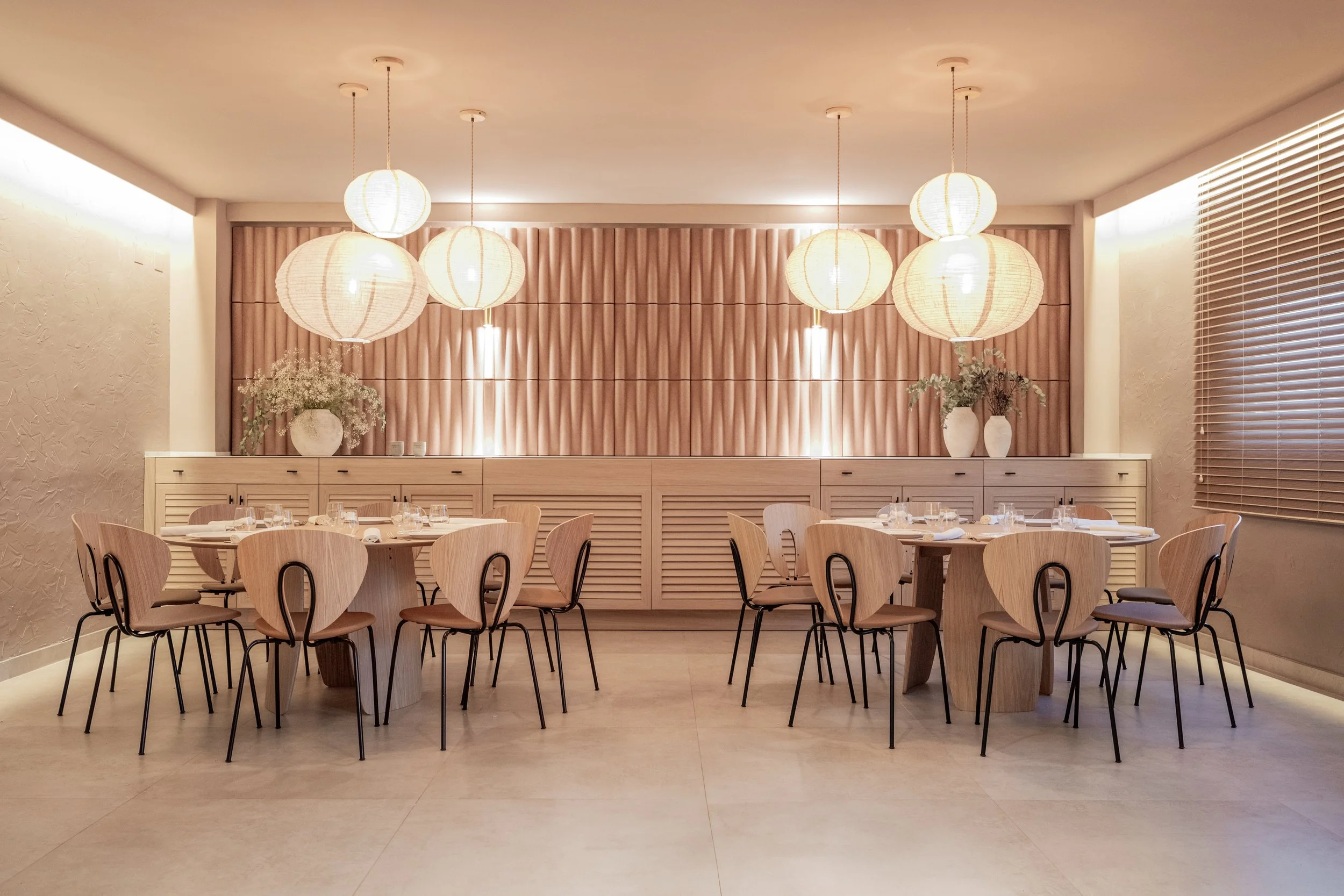 Sala de comedor elegante con mesas redondas, sillas de madera y decoraciones minimalistas, incluyendo lámparas de papel y jarrones con plantas, en tonos neutros y beige.