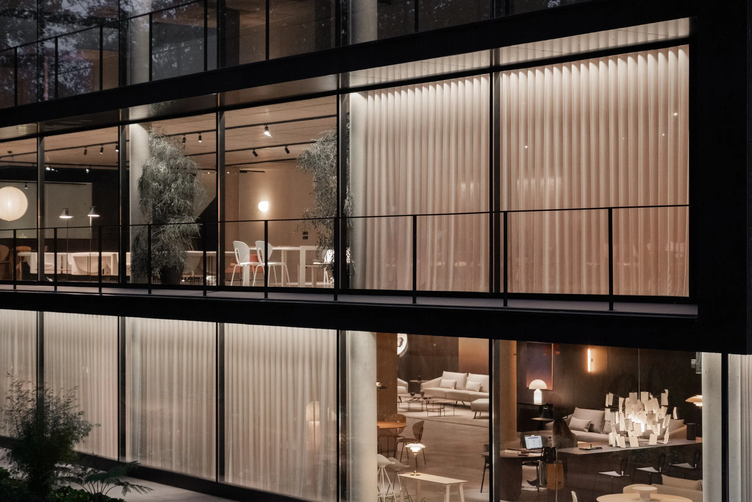 Vista de un edificio moderno con ventanas de vidrio que revelan un interior elegante y acogedor, decorado con muebles blancos y luz suave, en la noche.