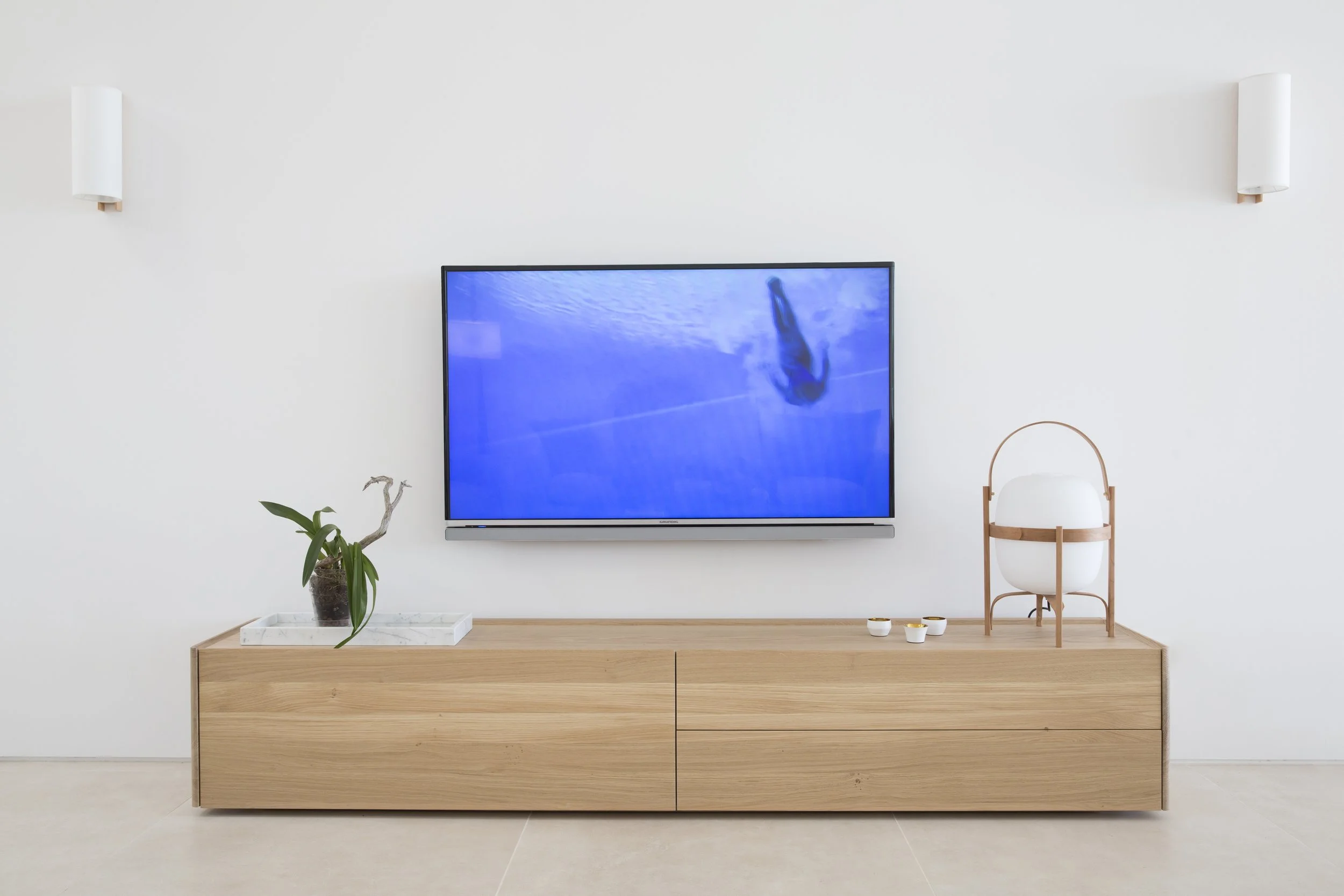 Mueble de madera con una televisión plana en la pared, una planta en una bandeja de mármol, y decoraciones blancas y doradas en un ambiente minimalista.
