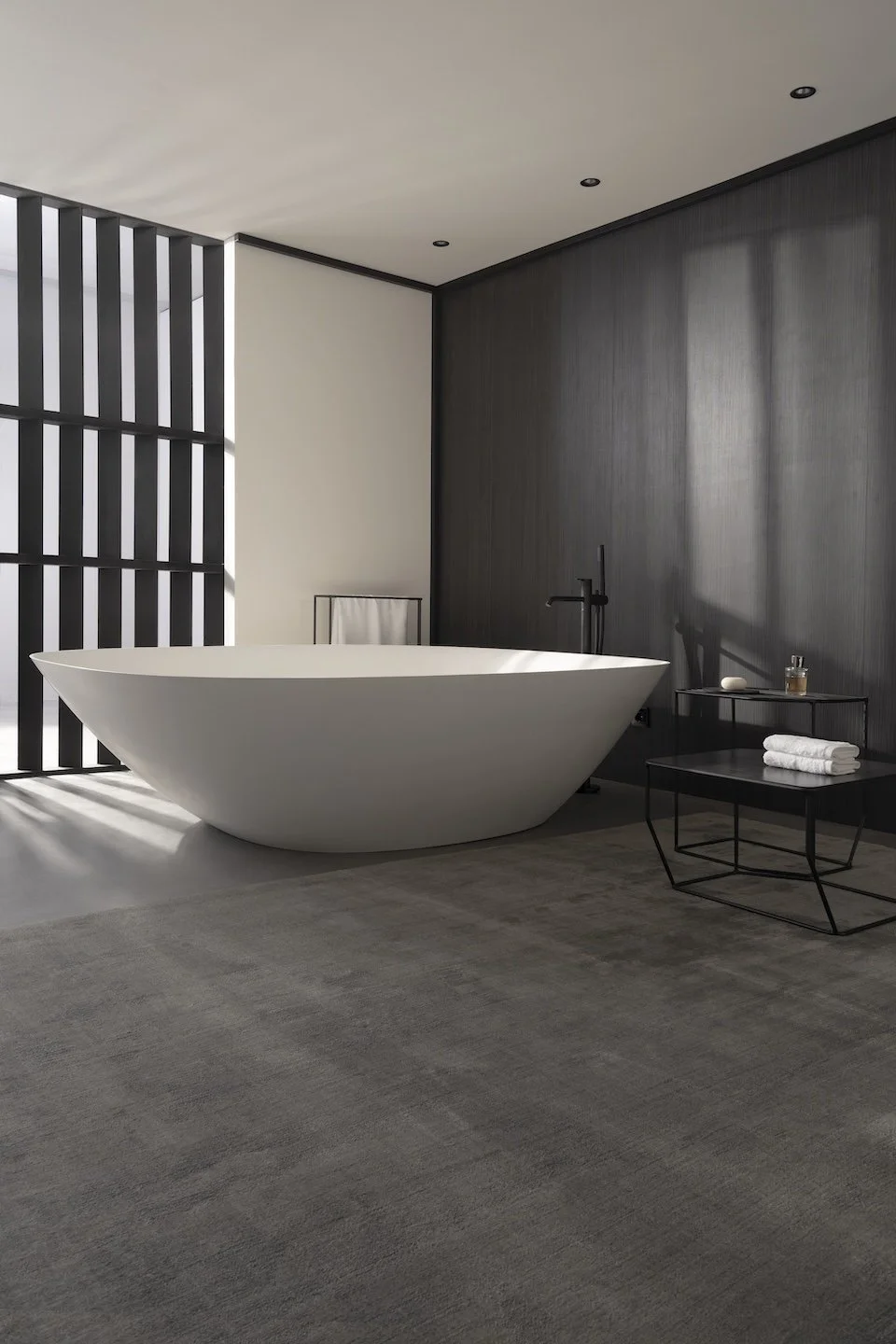 Baño moderno con bañera de pie blanca, paredes en tonos gris y negro, cortina de tela blanca, mesas para toallas y artículos de tocador, alfombra gris y luz natural entrando por una ventana con rejas negras.