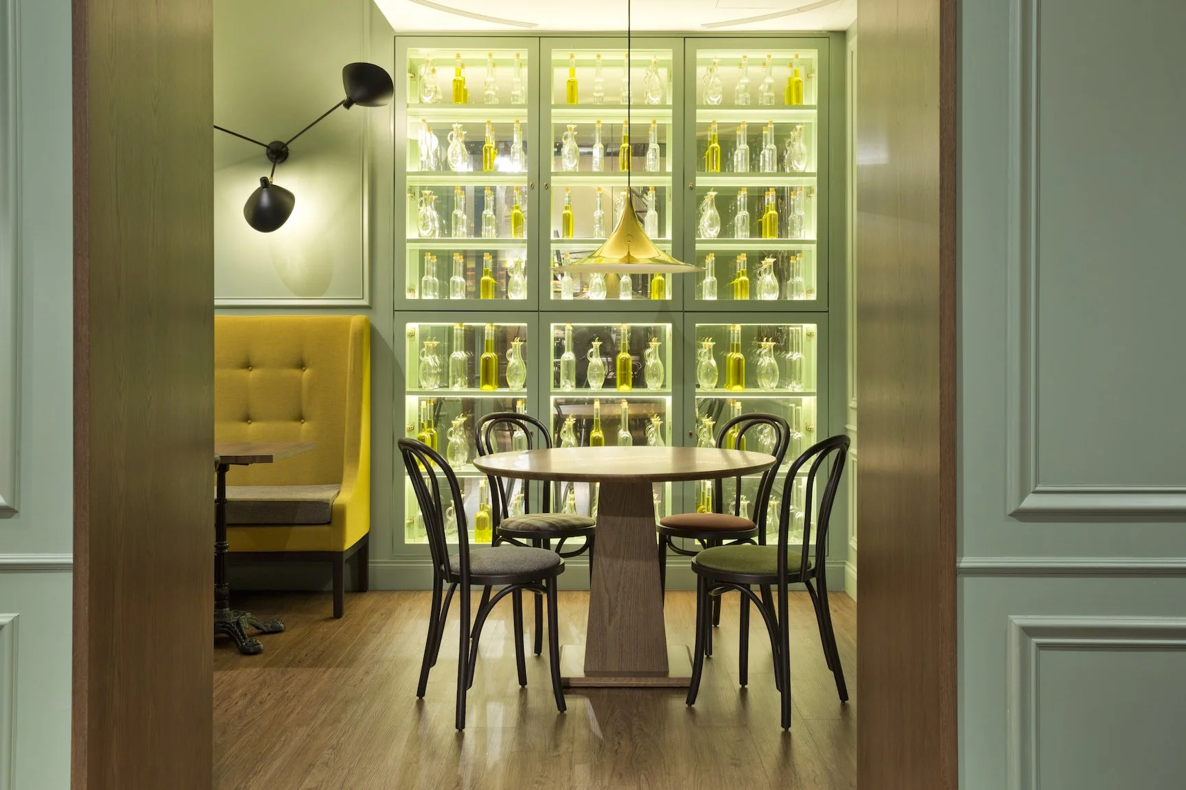 Sala de comedor con mesa redonda de madera y seis sillas, pared con vitrina iluminada que exhibe botellas de vino y decantadores, luminaria moderna y paredes en tonos pastel.