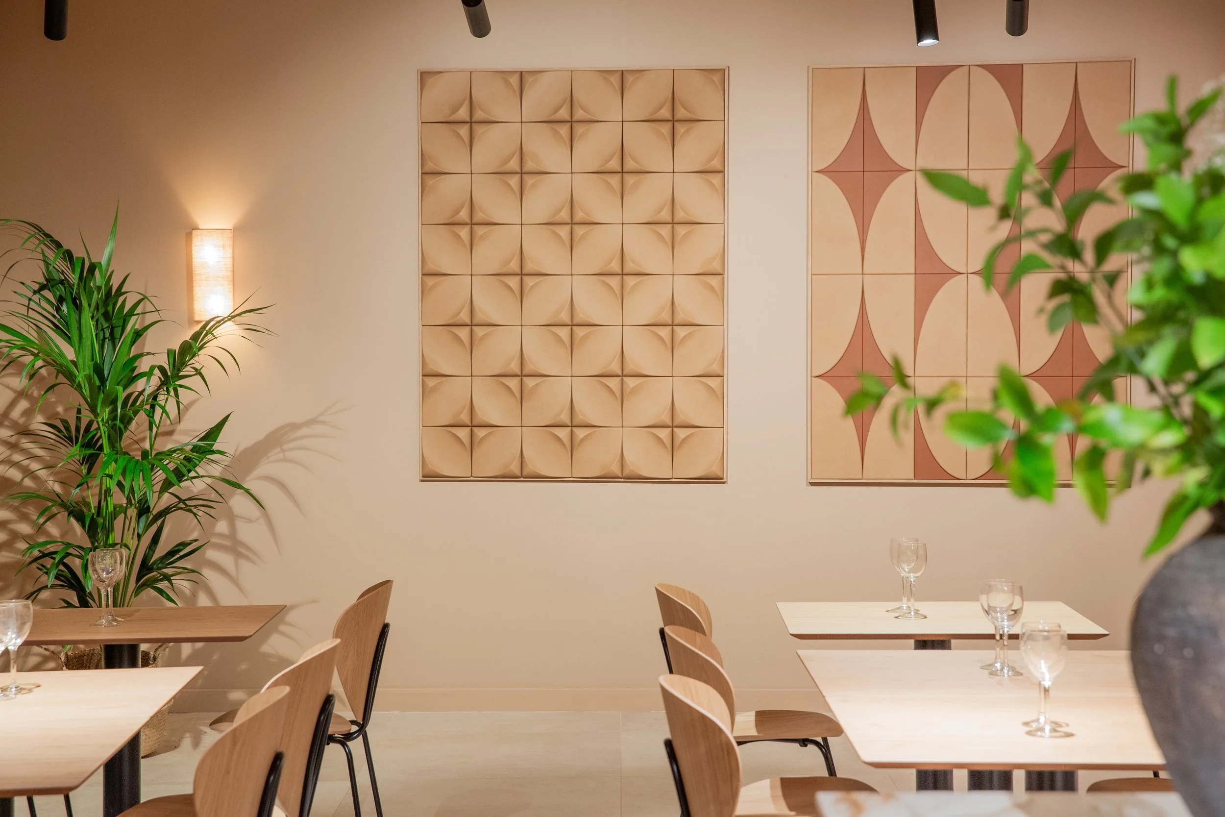 Interior de restaurante con mesas de madera, vasos de cristal, plantas verdes y obras de arte en la pared.