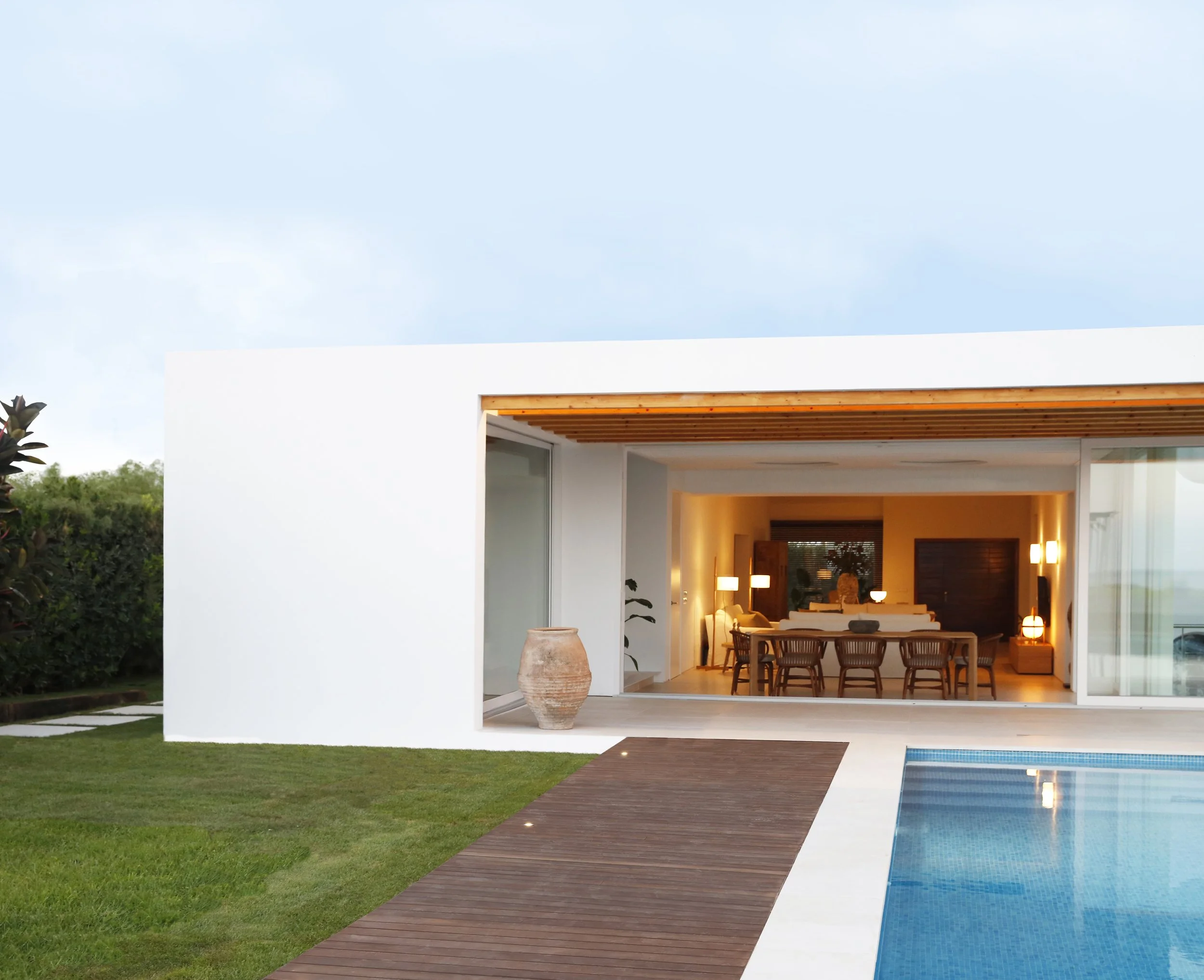 Vista frontal de una casa moderna con espacio interior visible, jardines, piscina y un cielo despejado.