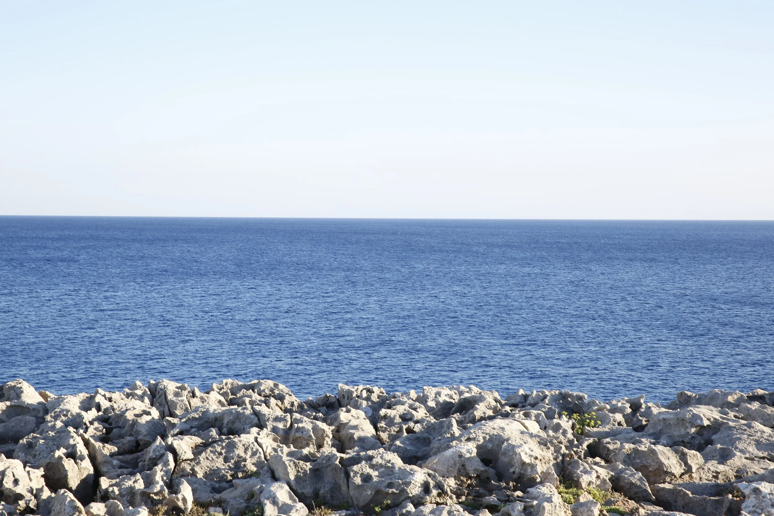 Vista del mar con aguas azules, rocas en primer plano y cielo despejado.