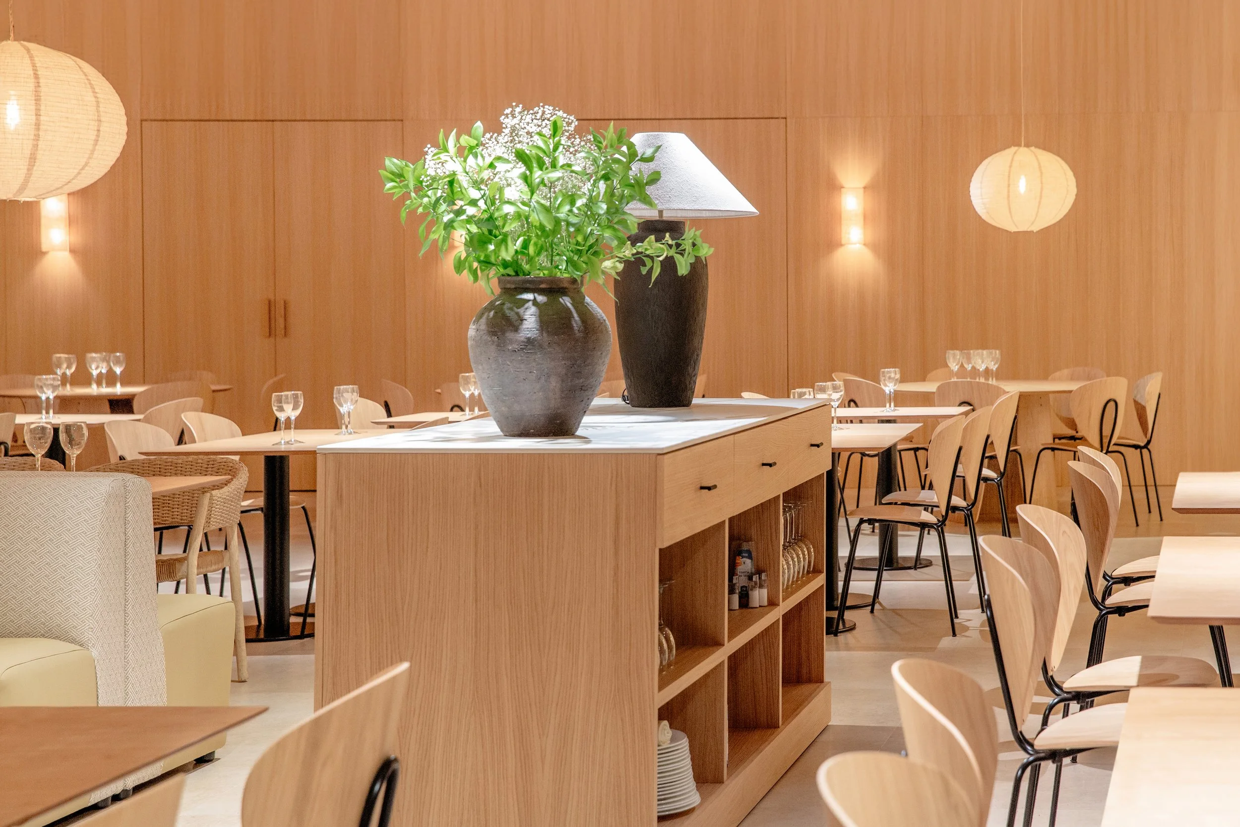 Interior de un restaurante con mesas y sillas de madera, decorado con un jarrón grande con plantas verdes y flores blancas en el centro de una mesa.