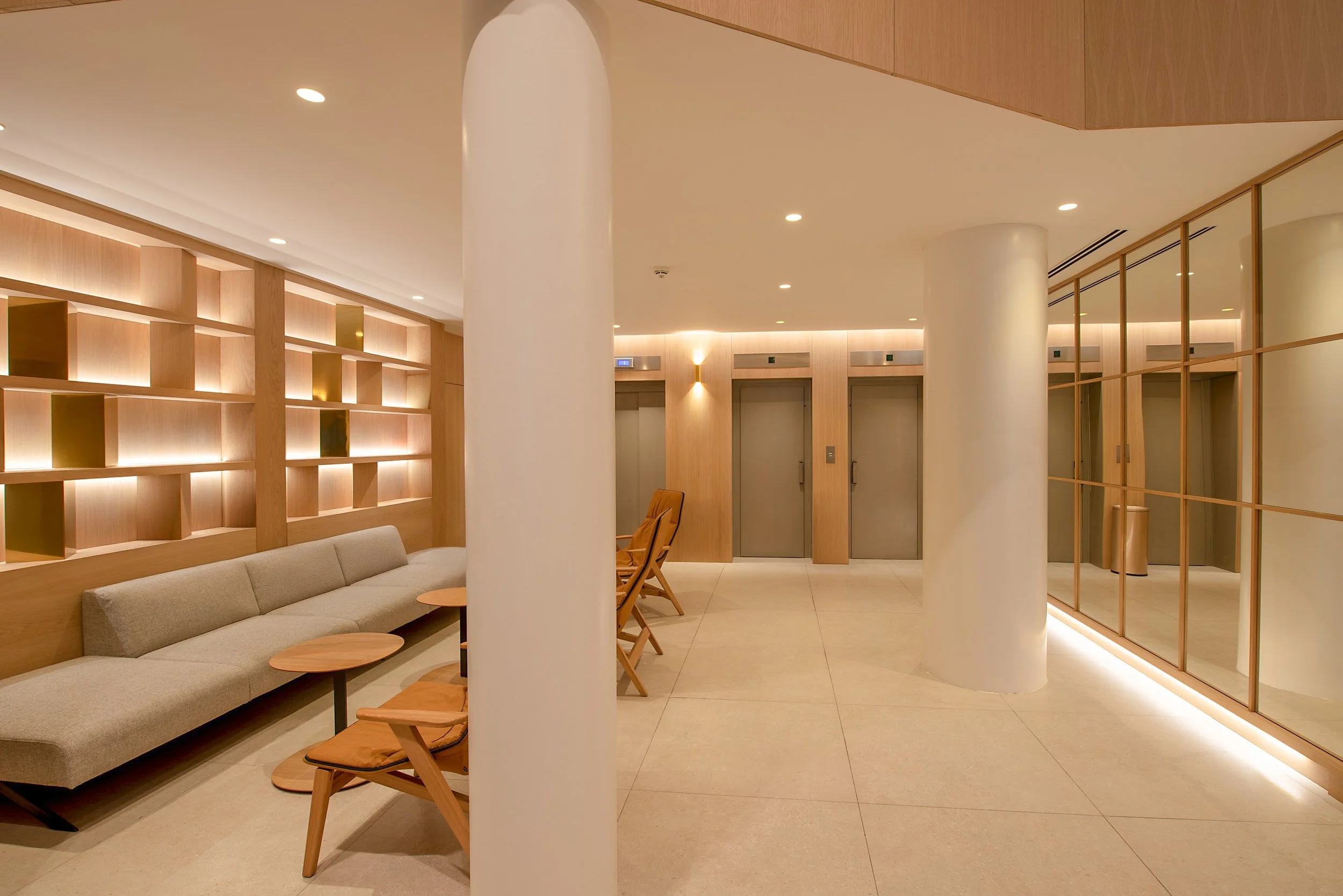 Sala de espera con paredes de madera, sillones grises y mesas pequeñas de madera, con puertas de ascensor y división de paneles de vidrio y madera en el fondo.