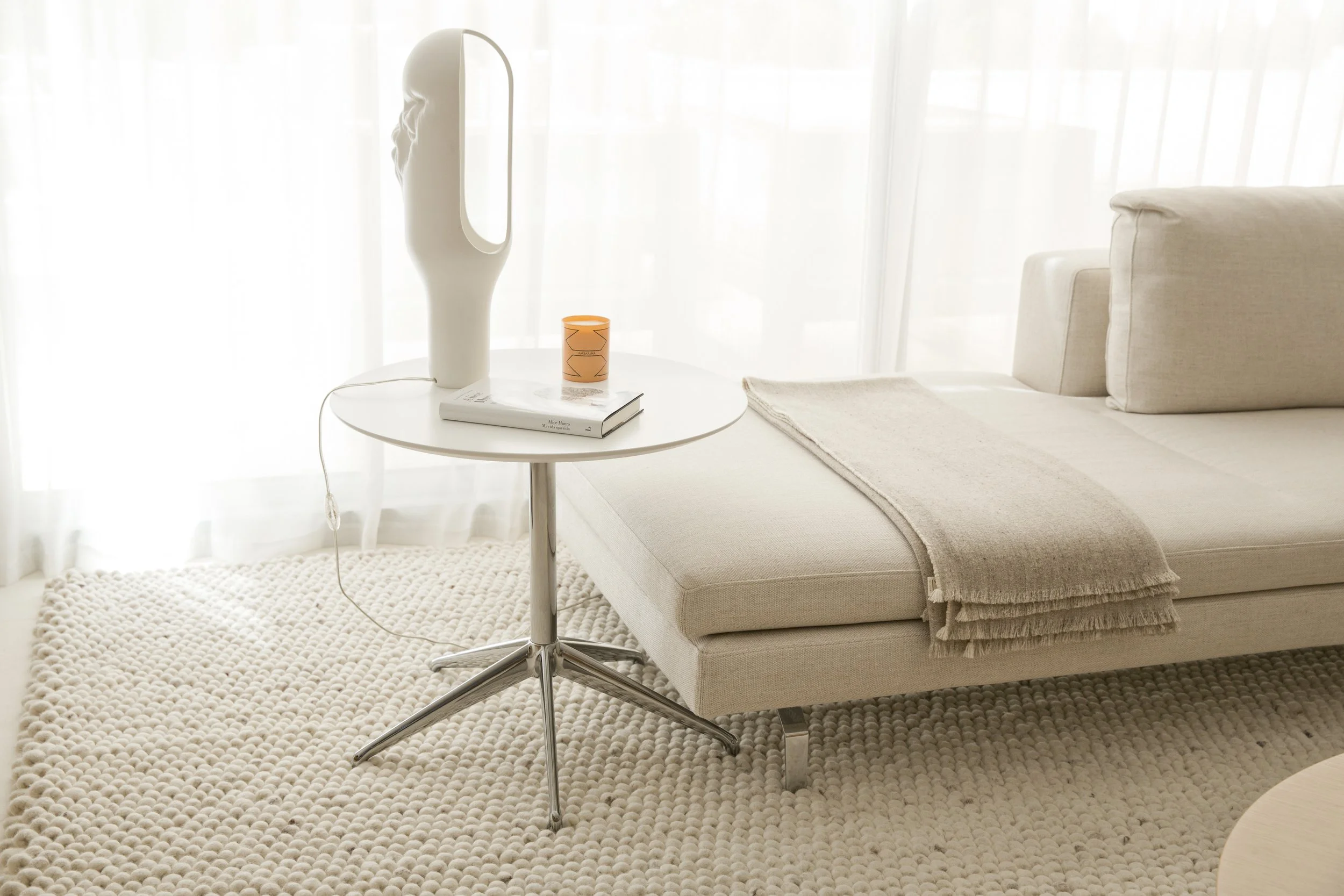 Sala de estar con sofá beige, mesa auxiliar blanca, lámpara moderna y una manta en el sofá, con cortinas blancas que dejan entrar luz natural.