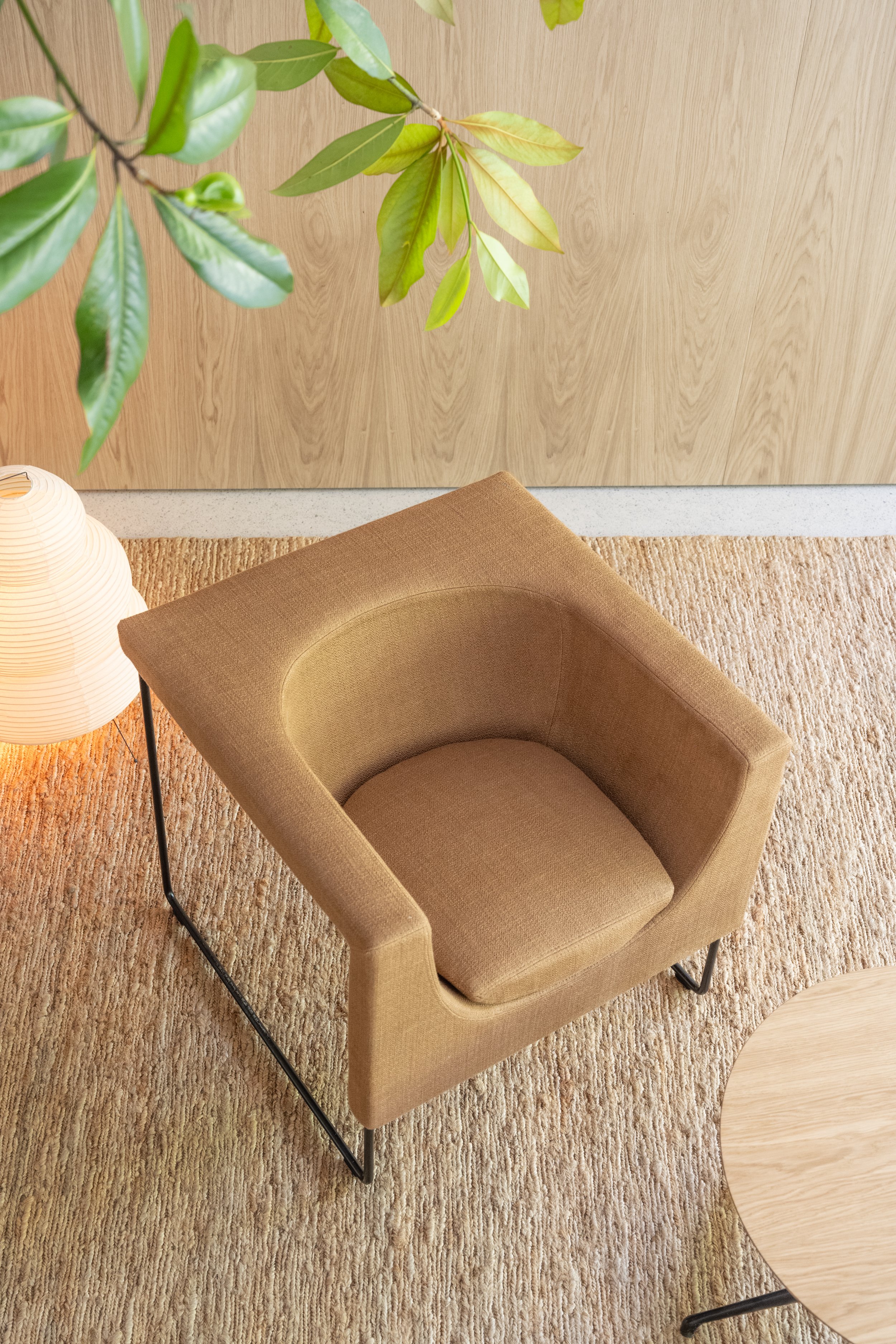 Silla de color beige en un espacio interior junto a una lámpara y una mesa redonda de madera clara, sobre una alfombra de color crema y con un fondo de pared de madera clara y plantas verdes.