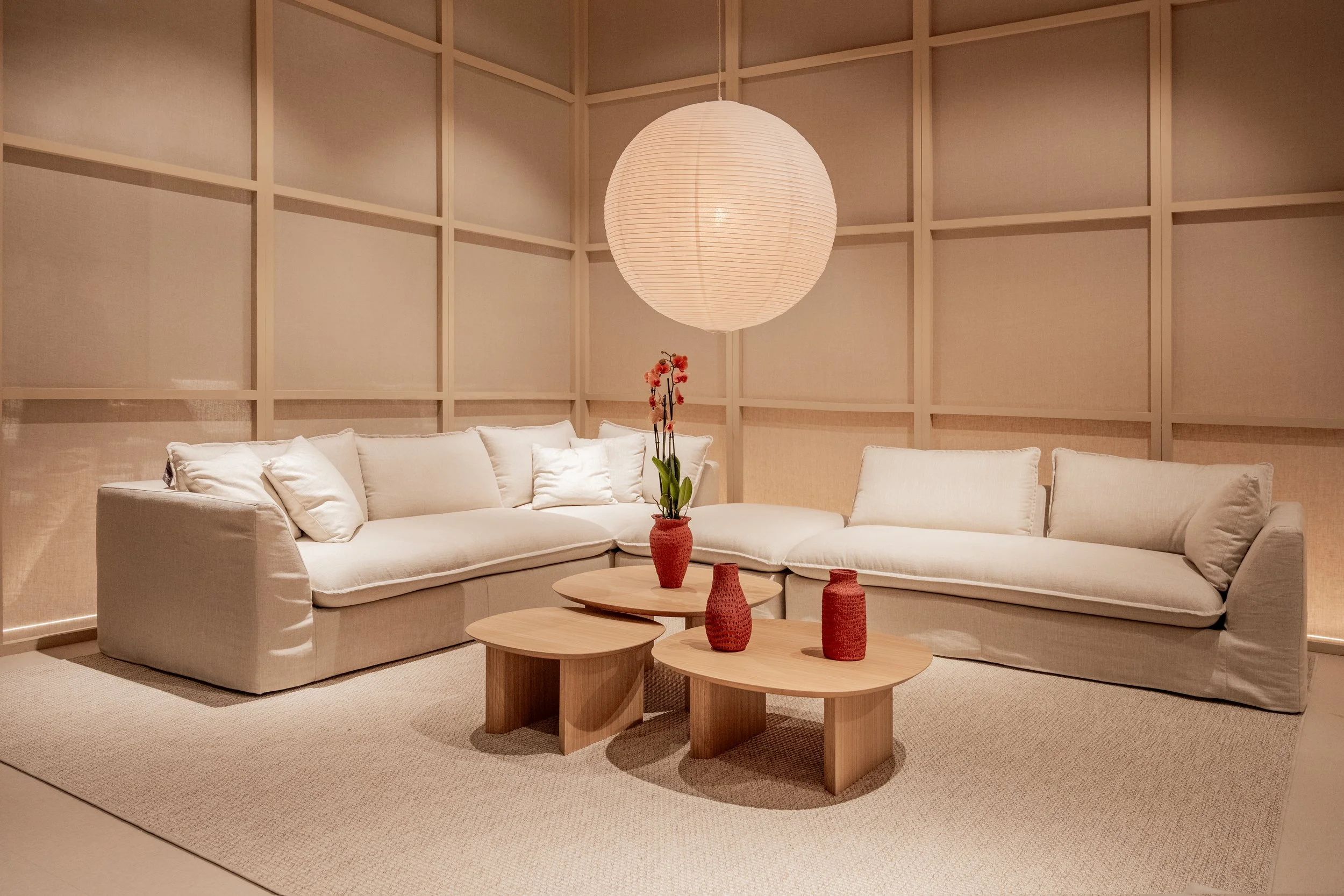 Sala de estar con un sofá de color crema, una mesa redonda de madera con jarrones rojos decorativos, una lámpara de papel en el techo y paredes revestidas con paneles de madera de tono claro.