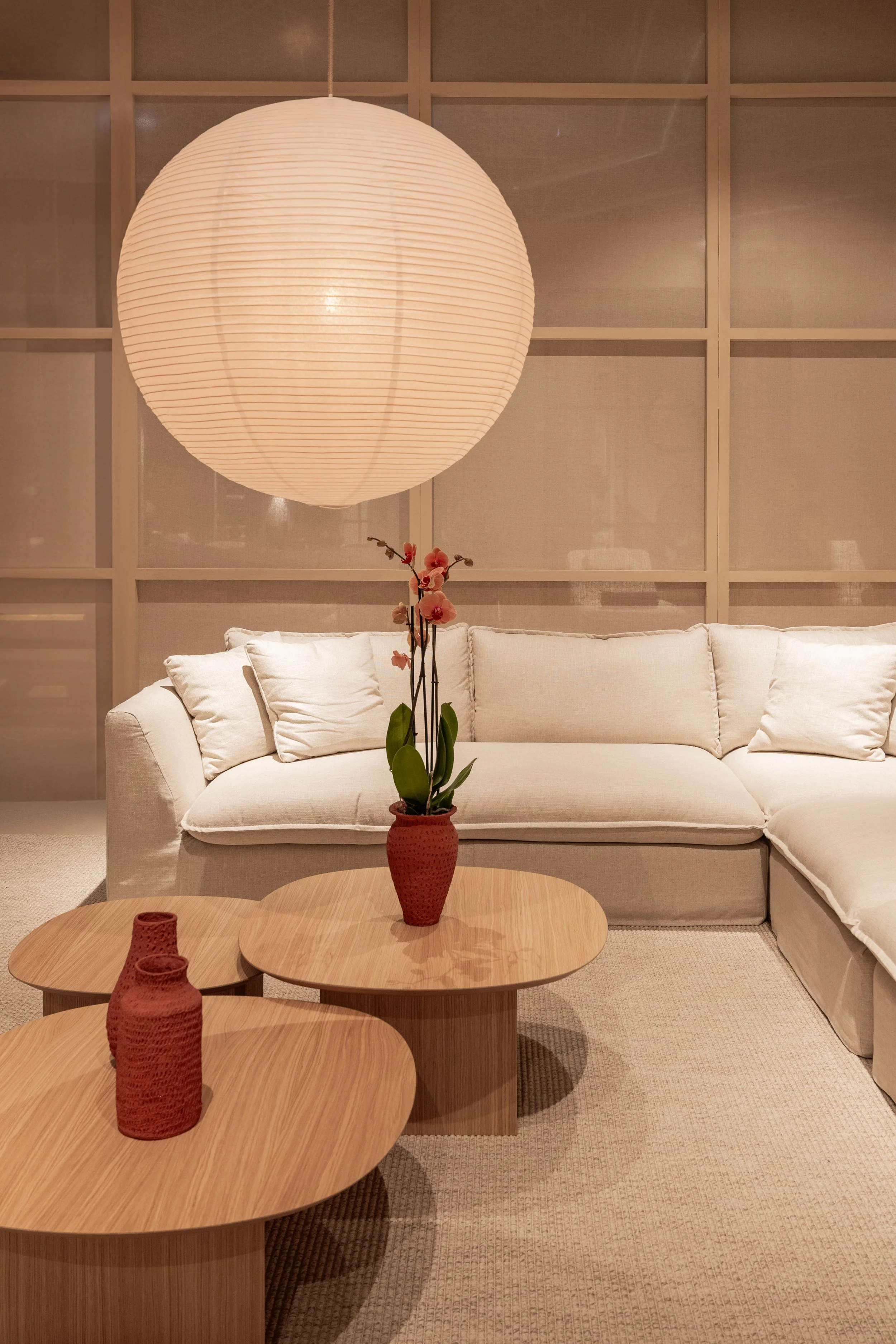 Sala con un sofá beige, una mesa de madera y jarrones rojos con flores de orquídea, lámpara de papel y paredes de paneles de madera.