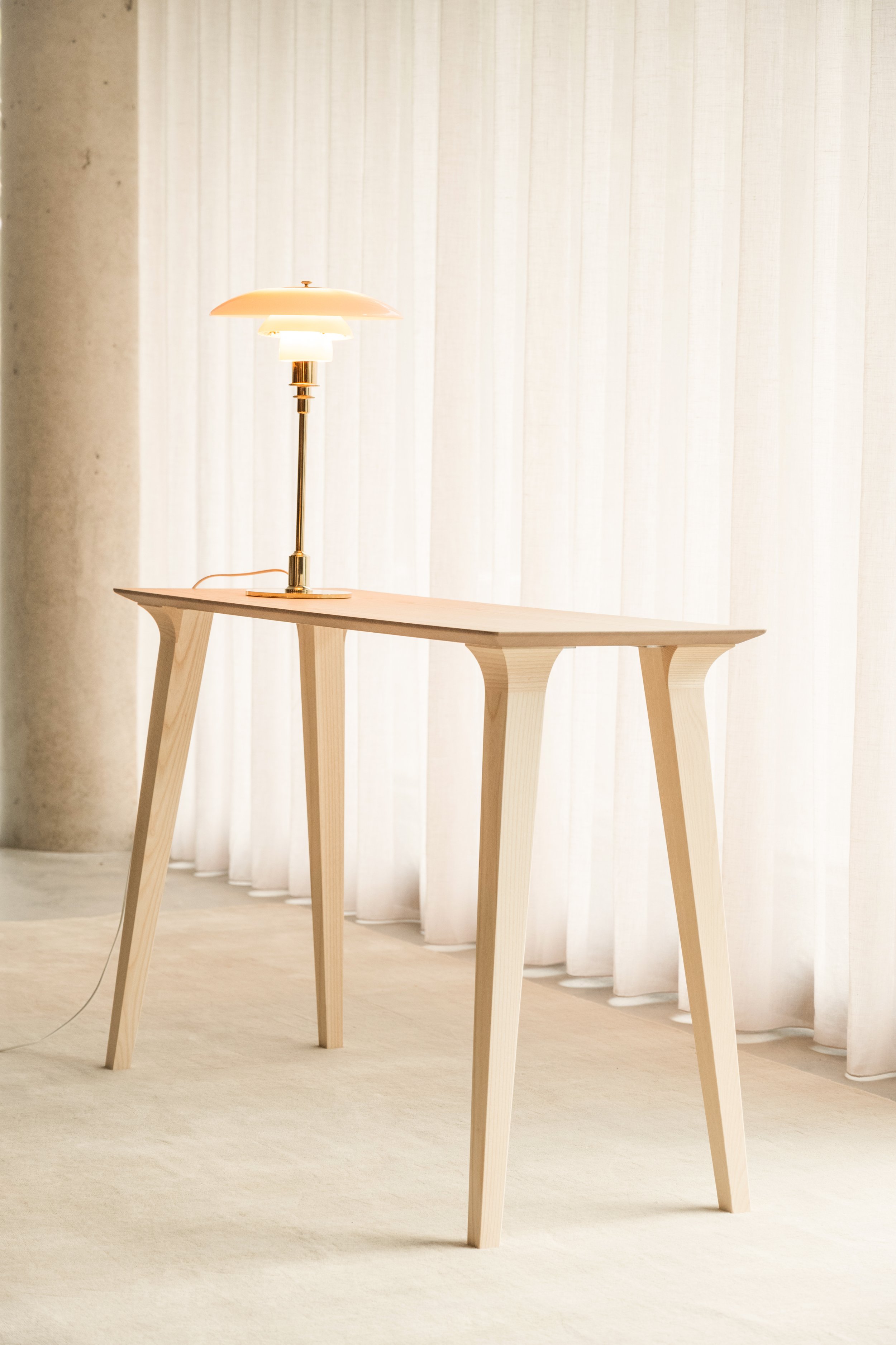 Mesa de madera con un lámpara moderna y blanca, decorada en un estilo minimalista, con cortinas blancas al fondo.