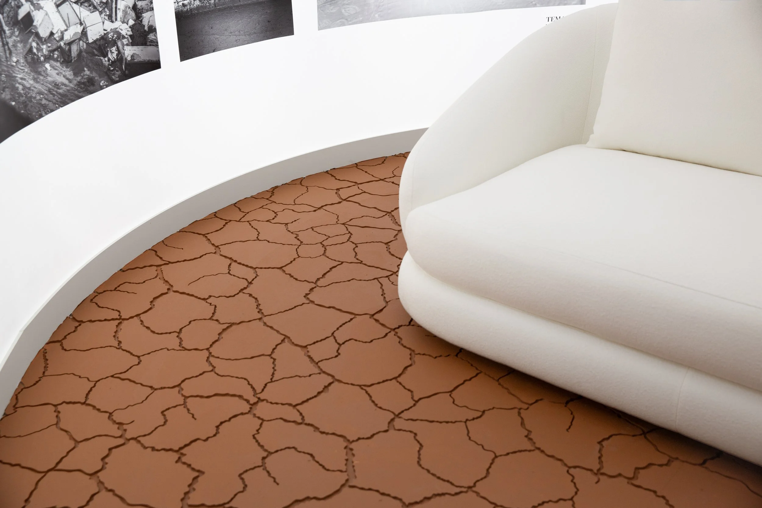 Vista de una sala con piso de barro con grietas en forma de red, pared blanca con fotografías enmarcadas, y un sillón blanco.