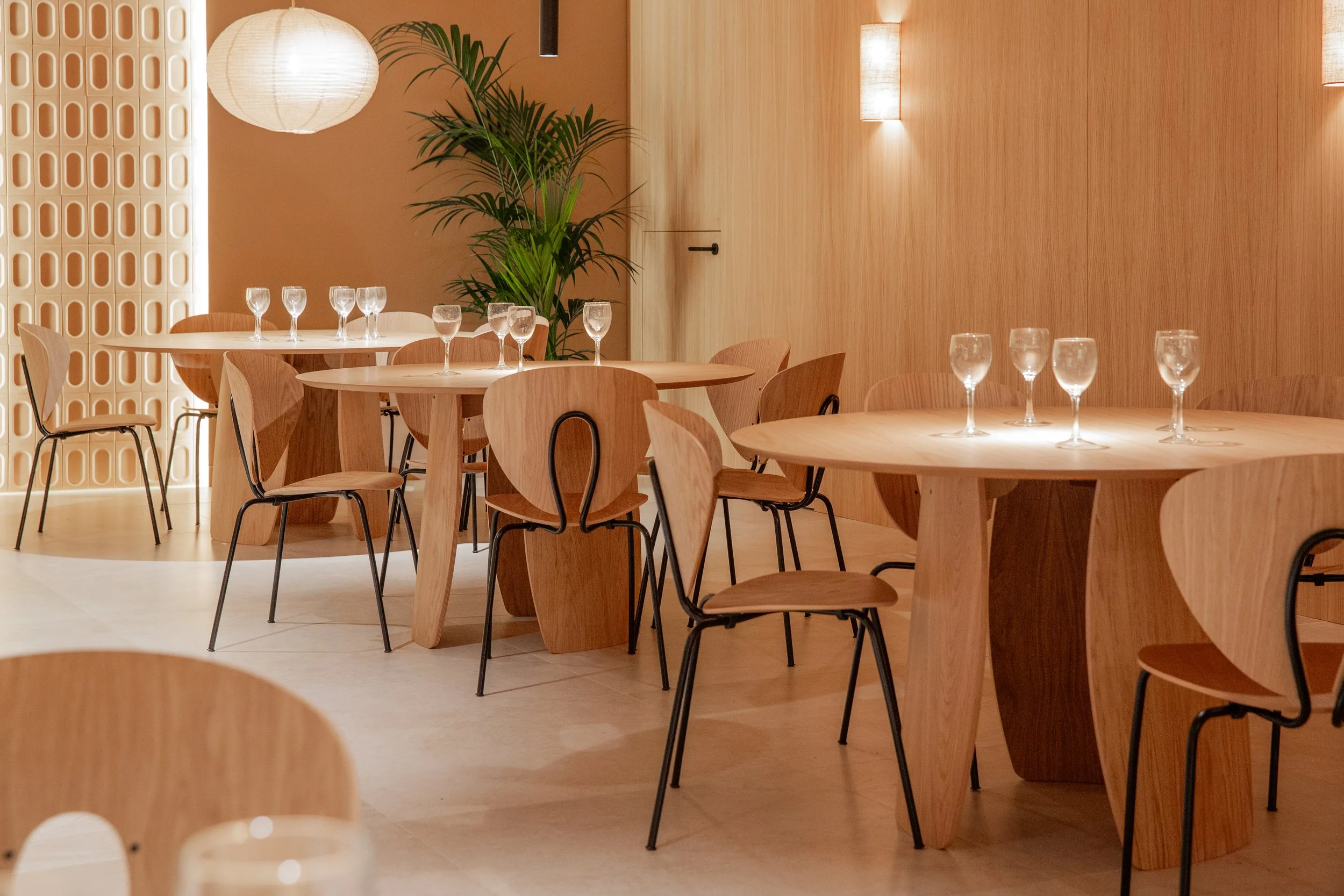 Sala de comedor con mesas de madera redondas, sillas de madera y metal, y copas de agua en las mesas. Decoración moderna con plantas verdes y paredes de madera clara, iluminación suave y lámparas en el techo.