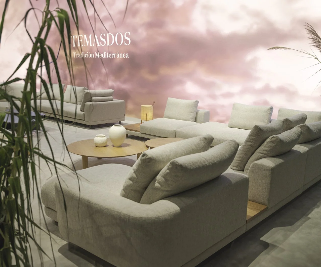 Sala de estar con sillones beige y mesas de madera, fondo de pared con imagen de cielo rosado y nubes, plantas verdes en primer plano, y texto que dice 'Temasdos, Tradición Mediterránea'.
