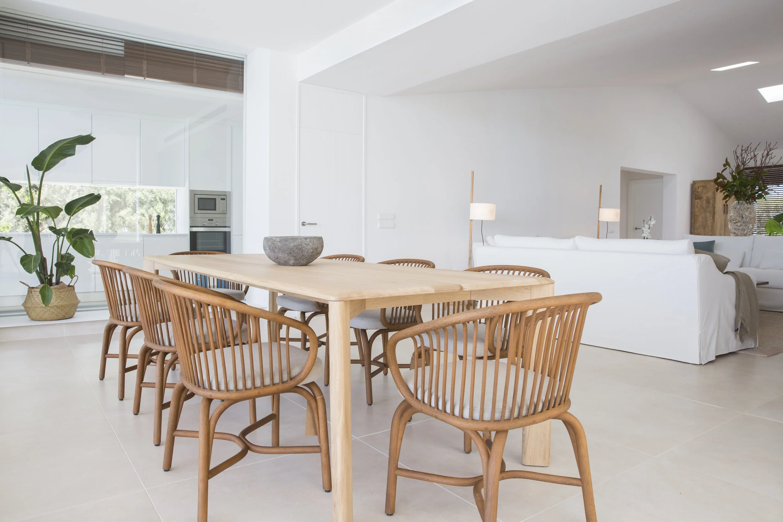 Interior de sala de estar moderna y luminosa con mesa de madera rodeada de sillas de madera y cojines en sofá blanco.